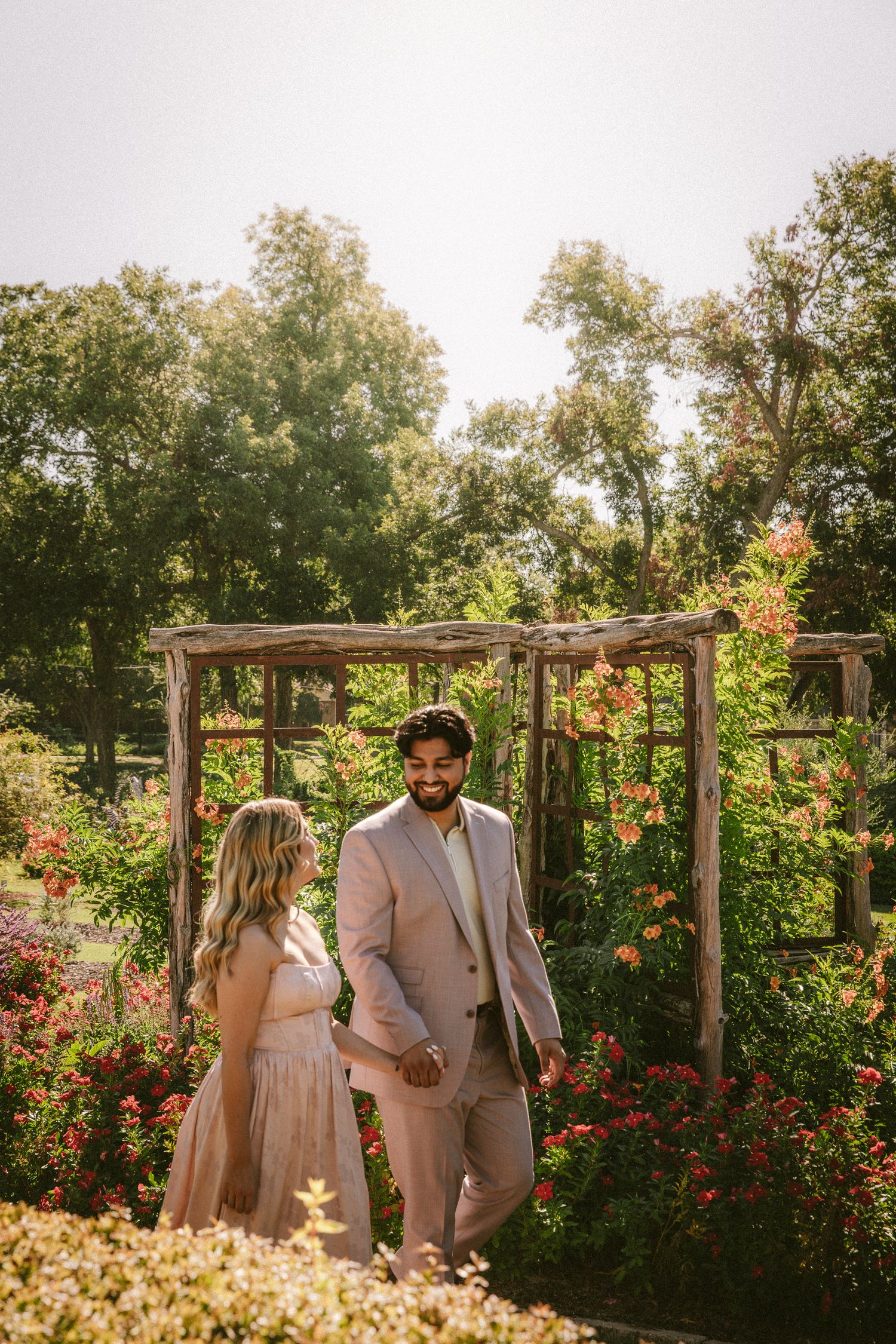 A couple poses together in a garden with wooden pergola and red roses in the background.