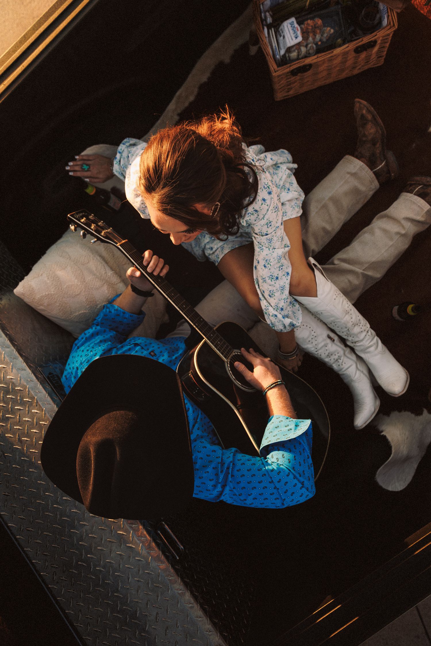 A person in a blue dress plays guitar while lying on a black leather couch in a dimly lit room.