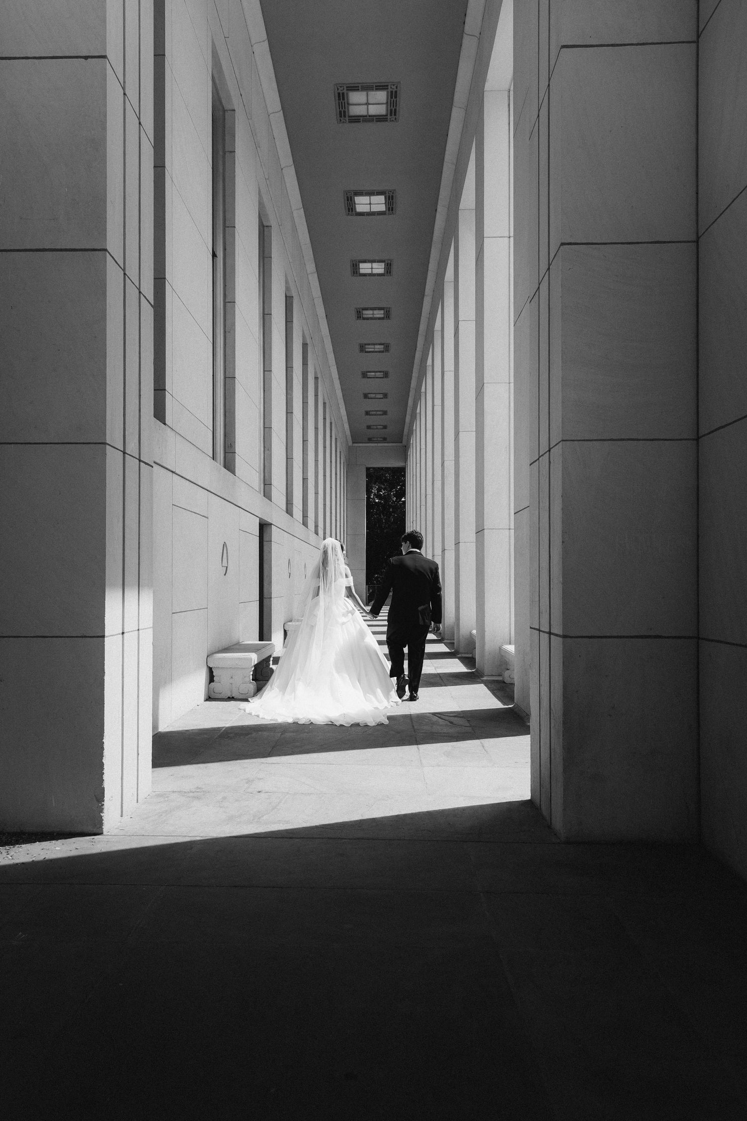 Bride in flowing white gown and groom walking down dramatic architectural corridor, black and white.