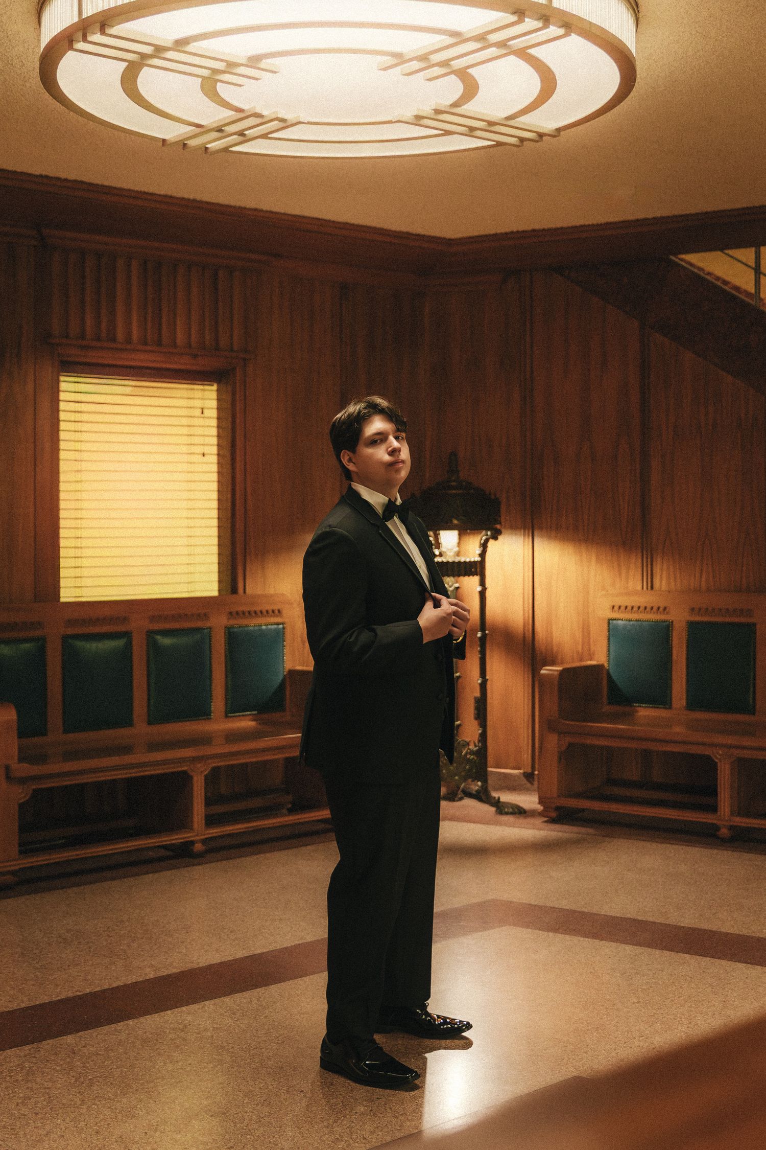 Groom in black tuxedo standing in warmly lit wood-paneled room with decorative ceiling light.