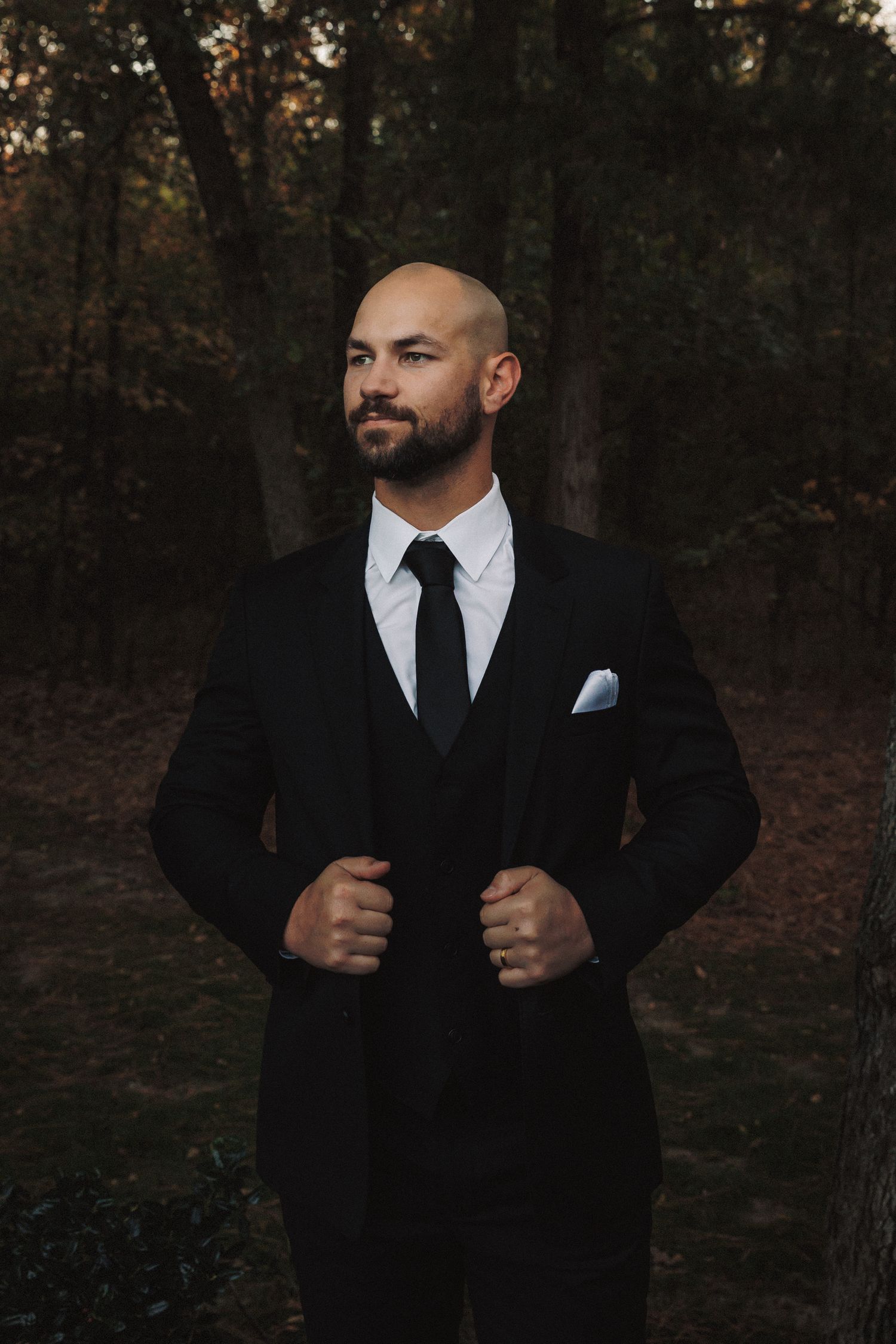 Confident bald groom in black suit with white pocket square poses in dark forest.