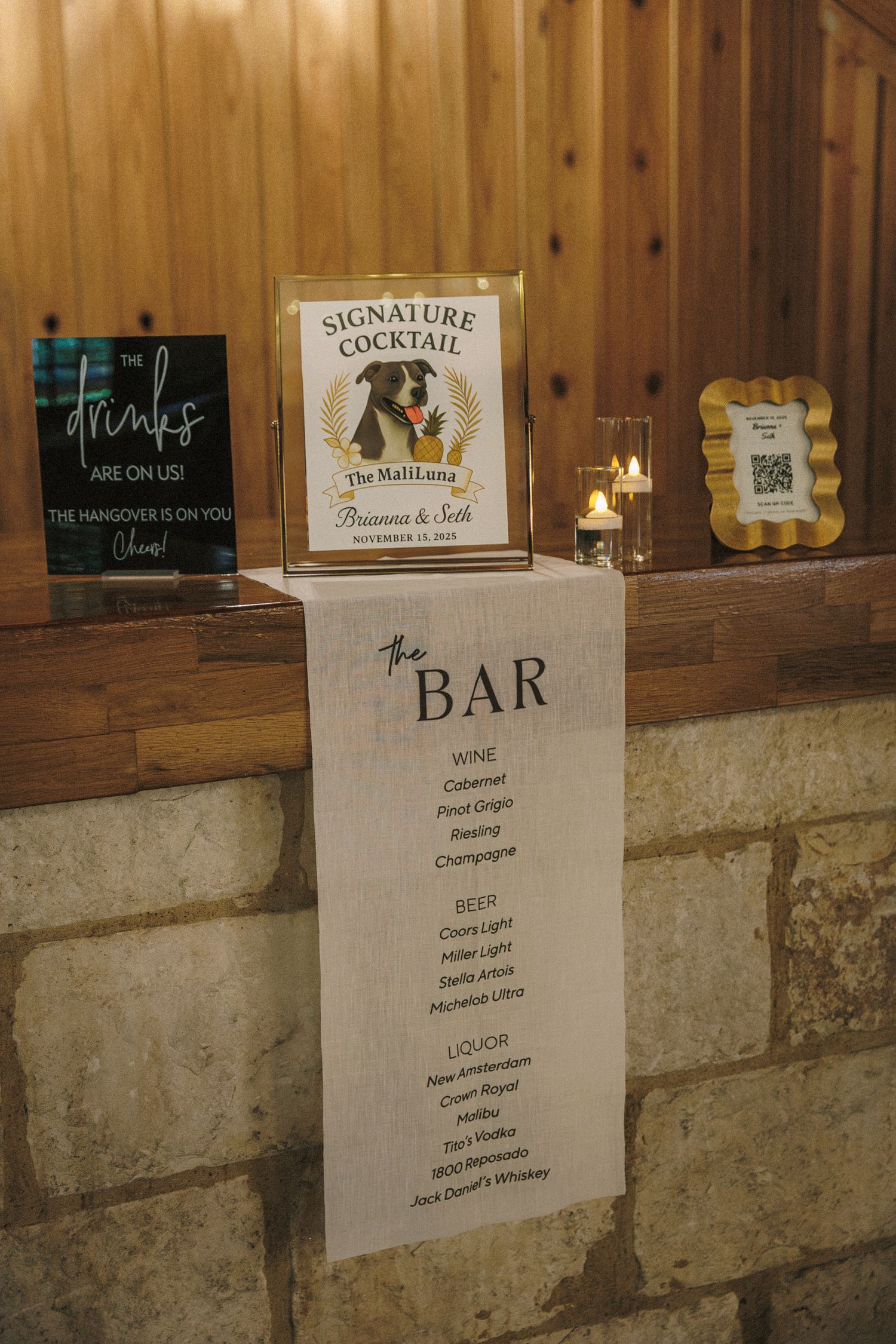 Wedding bar menu sign with signature cocktail display, candles, and drink options on rustic stone wall.