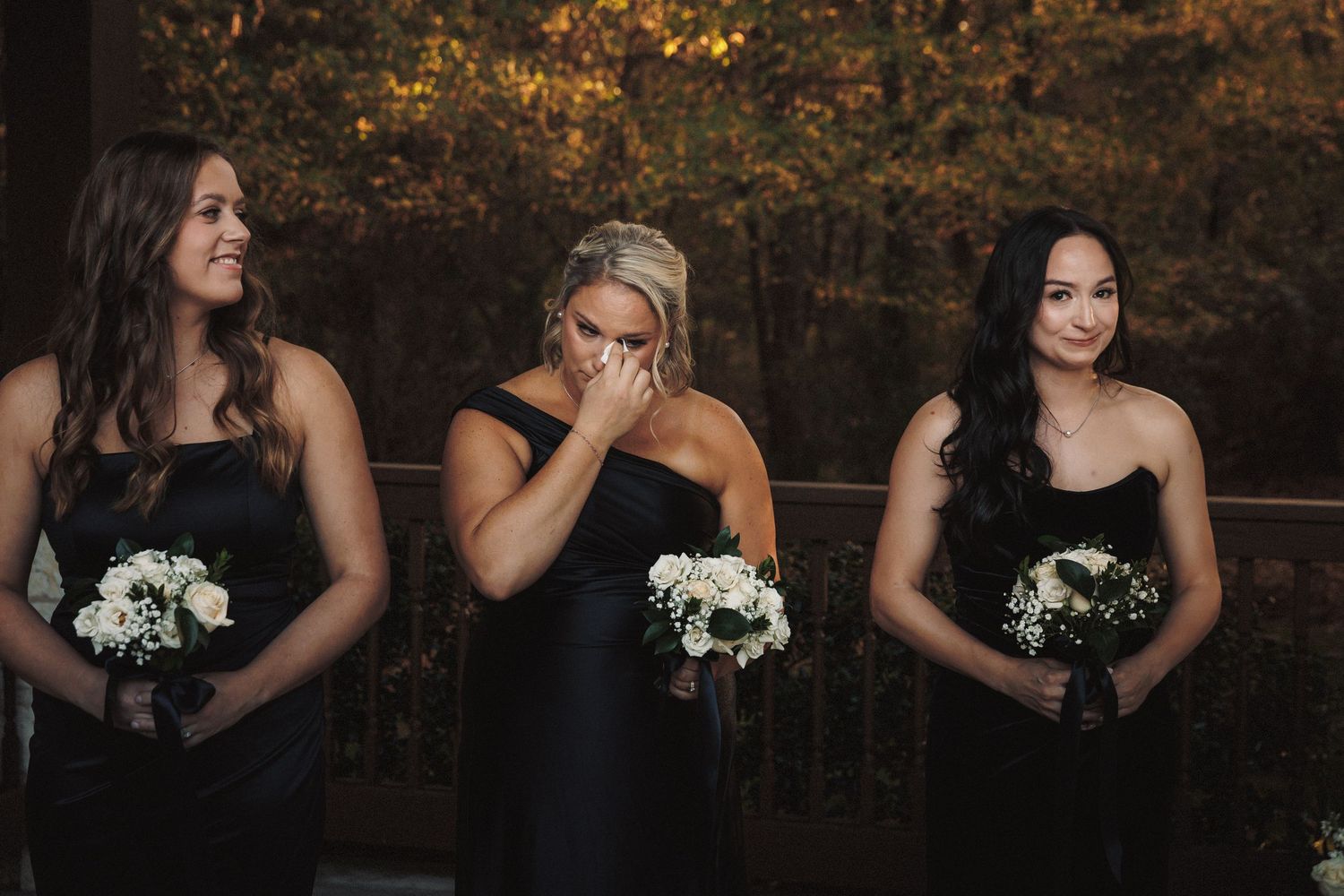Three bridesmaids in black dresses holding white bouquets with one wiping tears at a wedding.