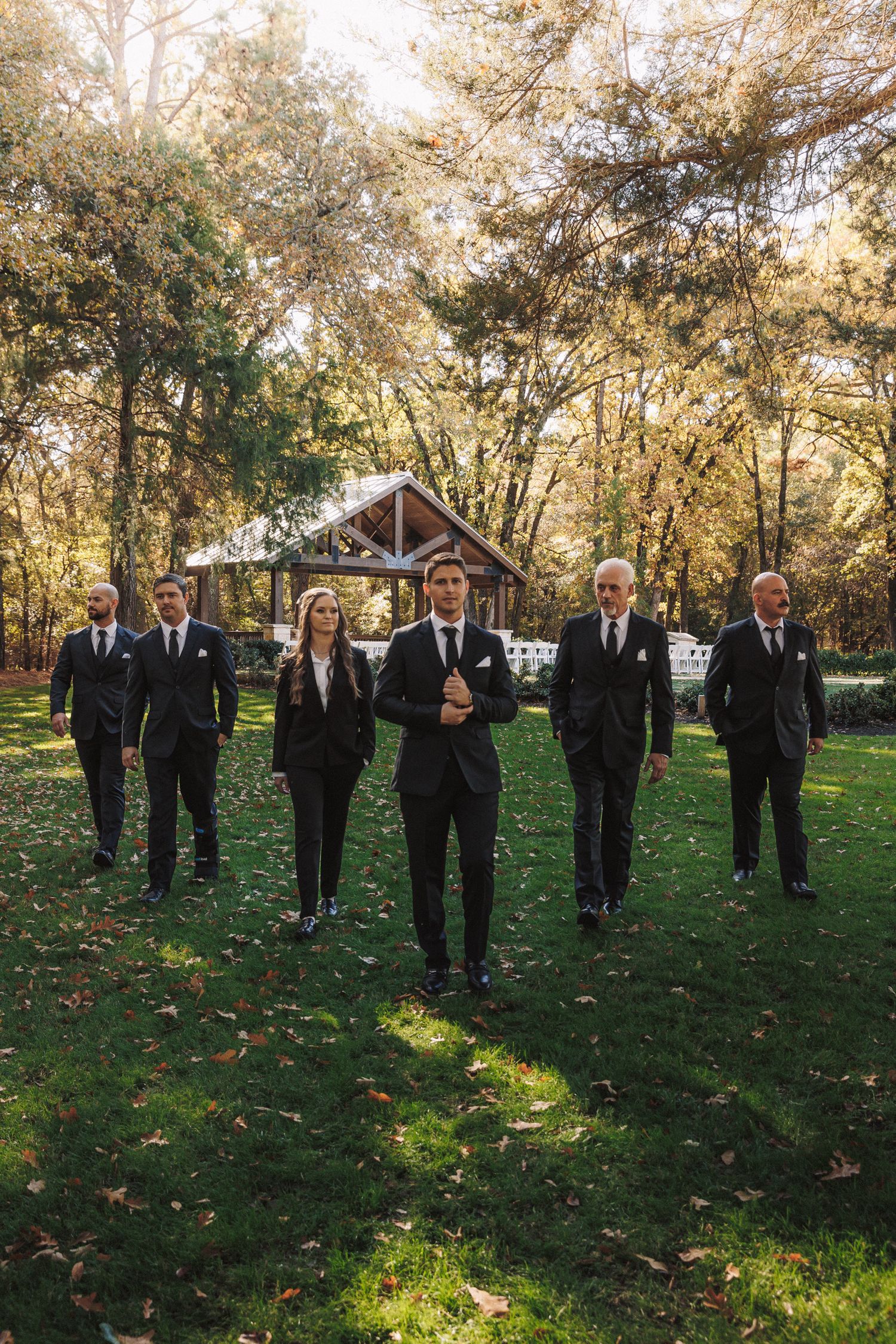 Groomsmen in black suits walking outdoors at a woodland wedding venue with a pavilion in the background.