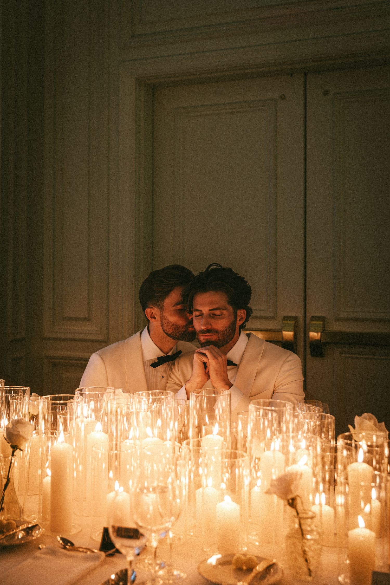 Two grooms in white tuxedos sit together surrounded by glowing candles at their wedding reception.