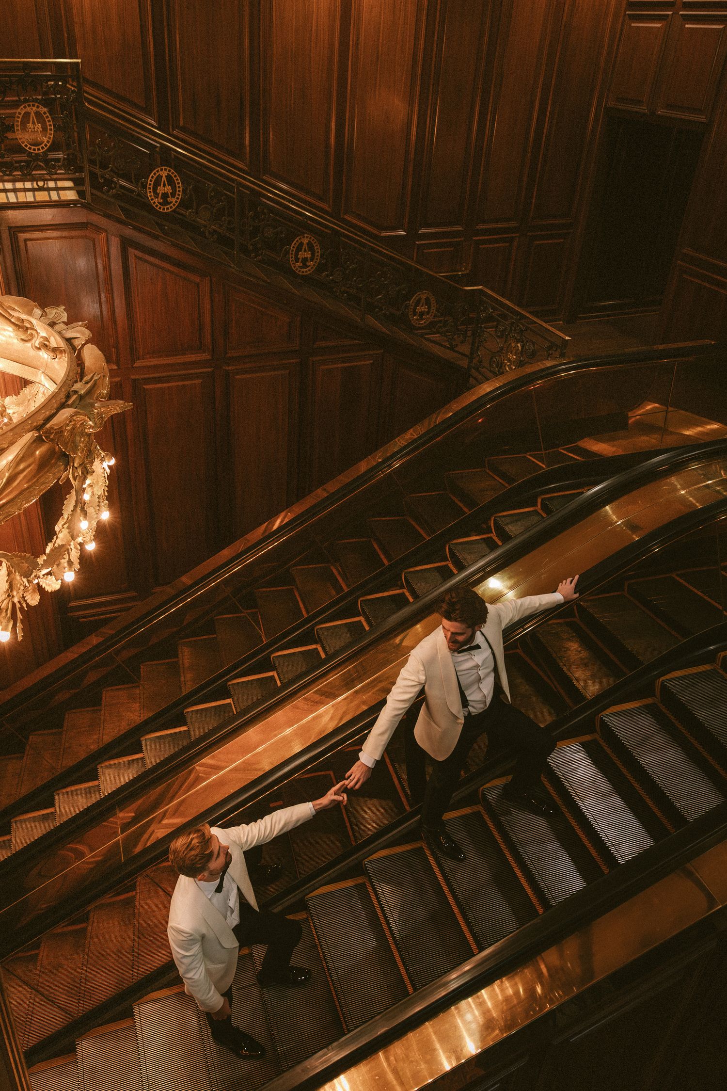 Aerial view of two grooms in white suits holding hands on an ornate wooden escalator in elegant venue.