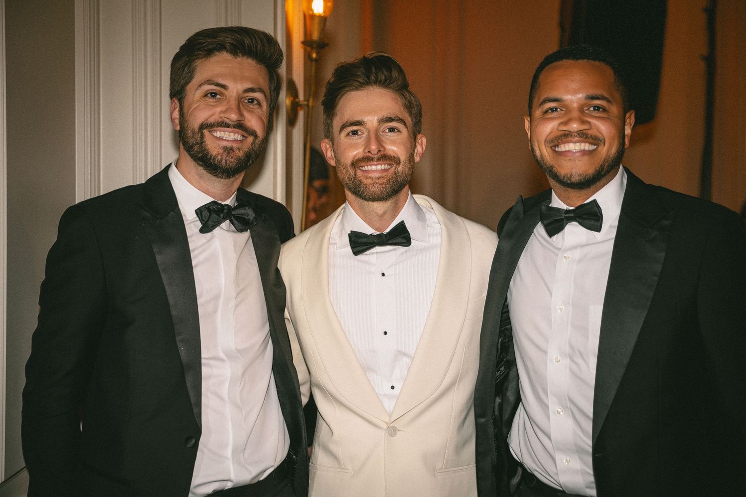 Three smiling men in tuxedos pose together at a wedding celebration.