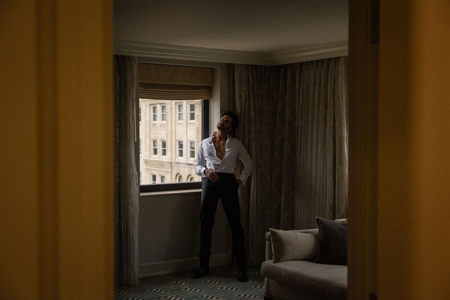 Groom in white shirt talking on phone by hotel window with city view.