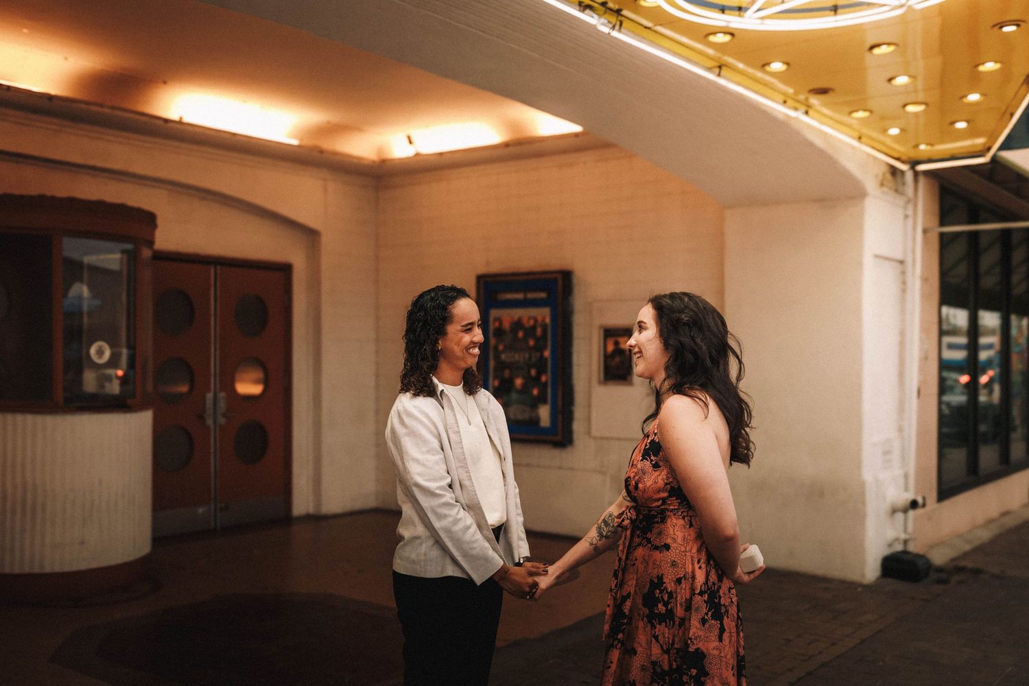 Two women holding hands and smiling at each other outside a vintage theater at night.