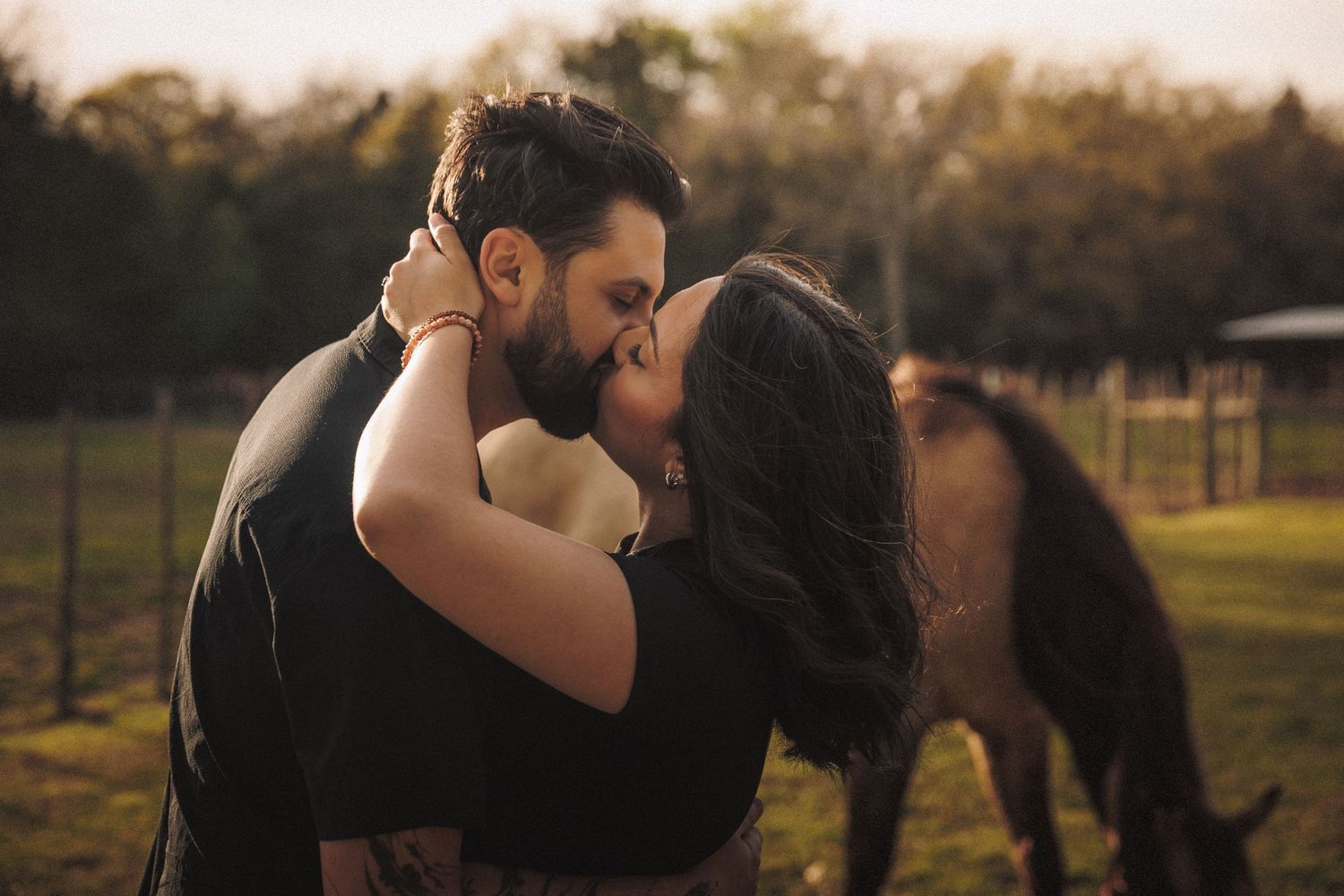 Couple sharing a romantic kiss on a farm with a horse grazing in the background.