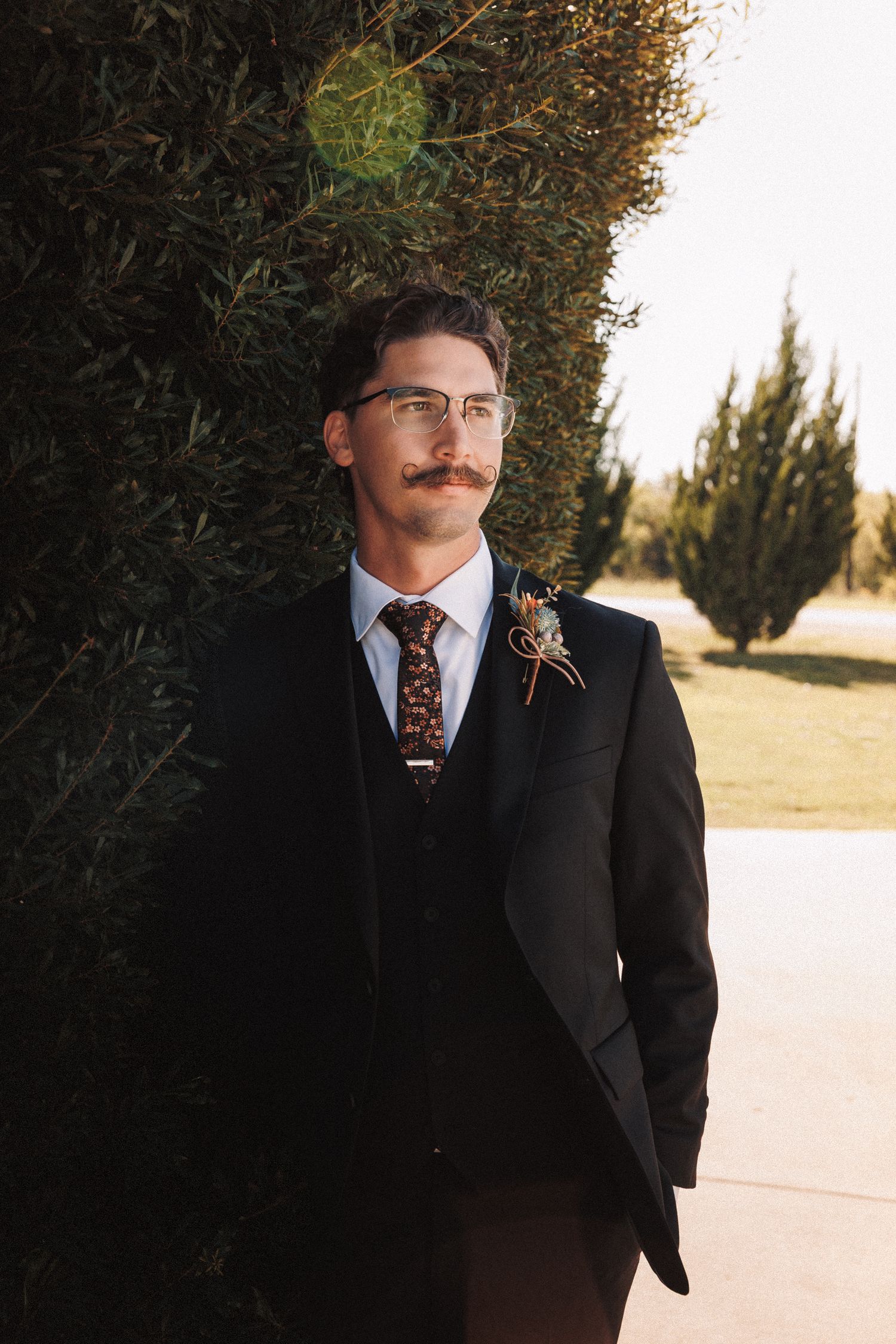 Dapper groom with mustache and glasses wearing a black suit with floral boutonniere outdoors.