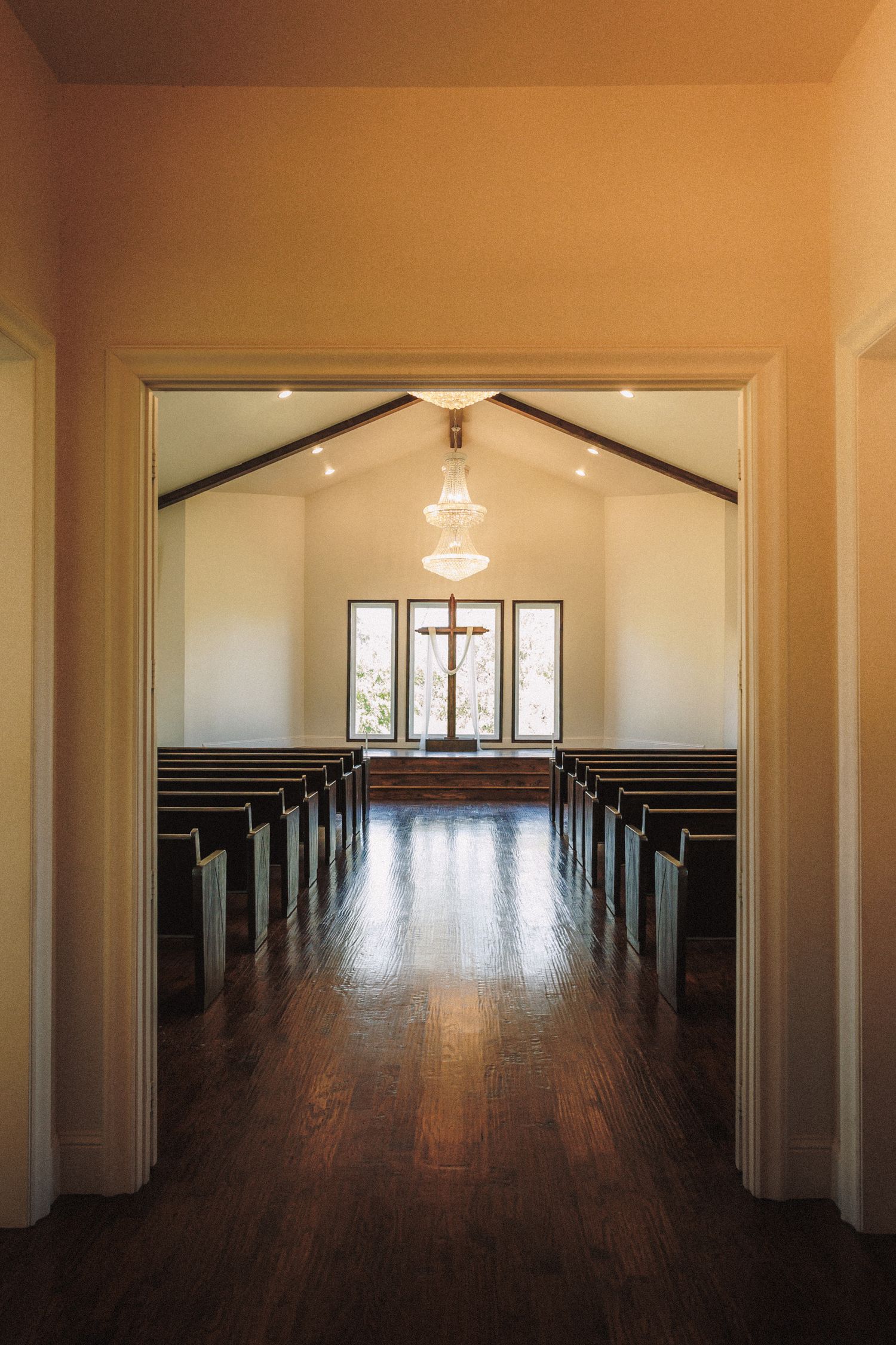 Serene chapel interior with wooden pews, cross, chandelier, and hardwood floors.
