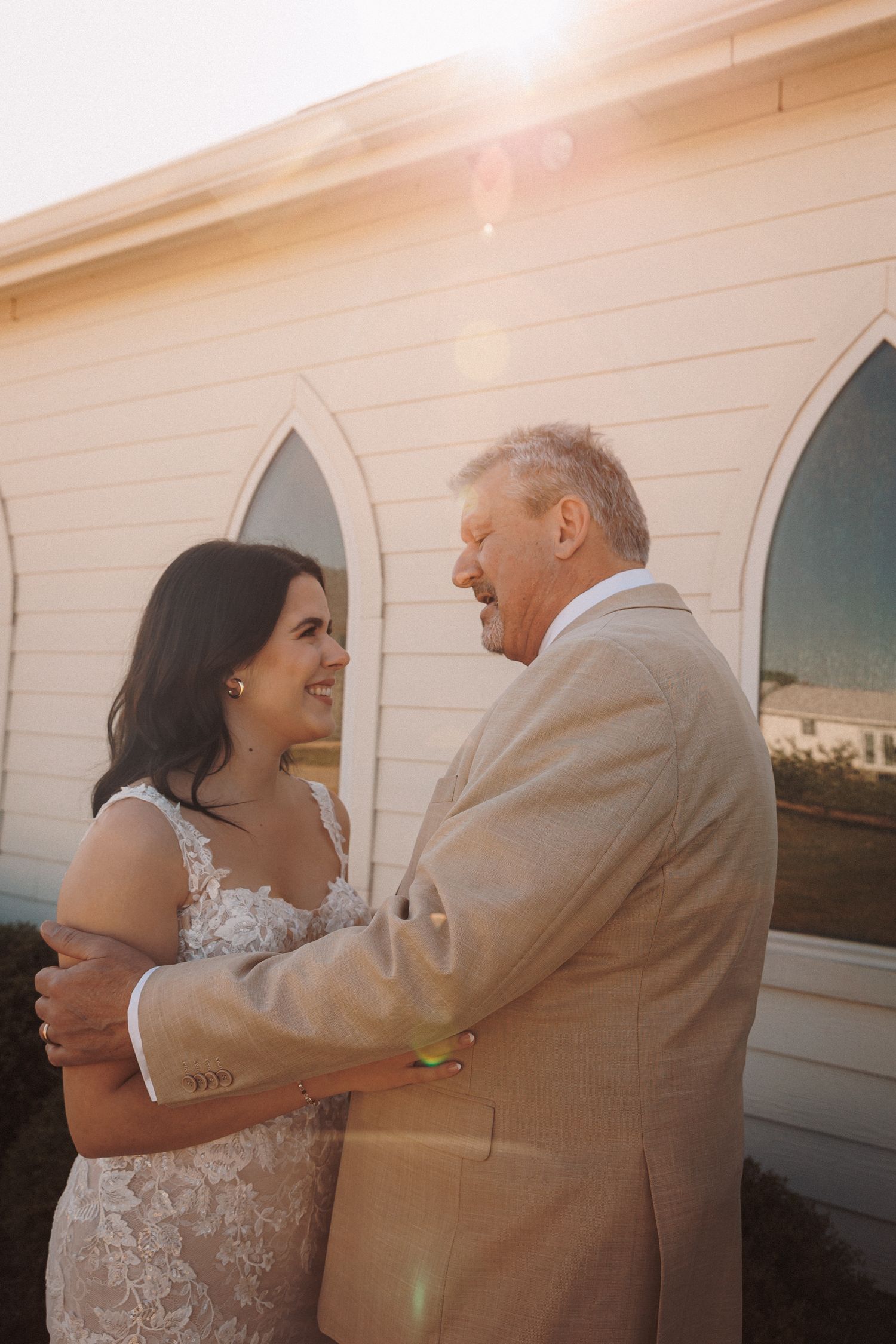 Bride and older man in tan suit embrace and smile outside a white chapel during golden hour sunlight.