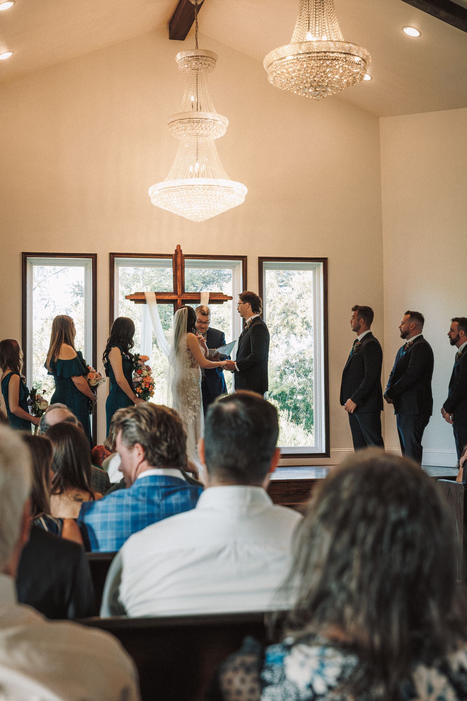 Wedding ceremony inside chapel with chandelier, cross, and guests seated in pews.
