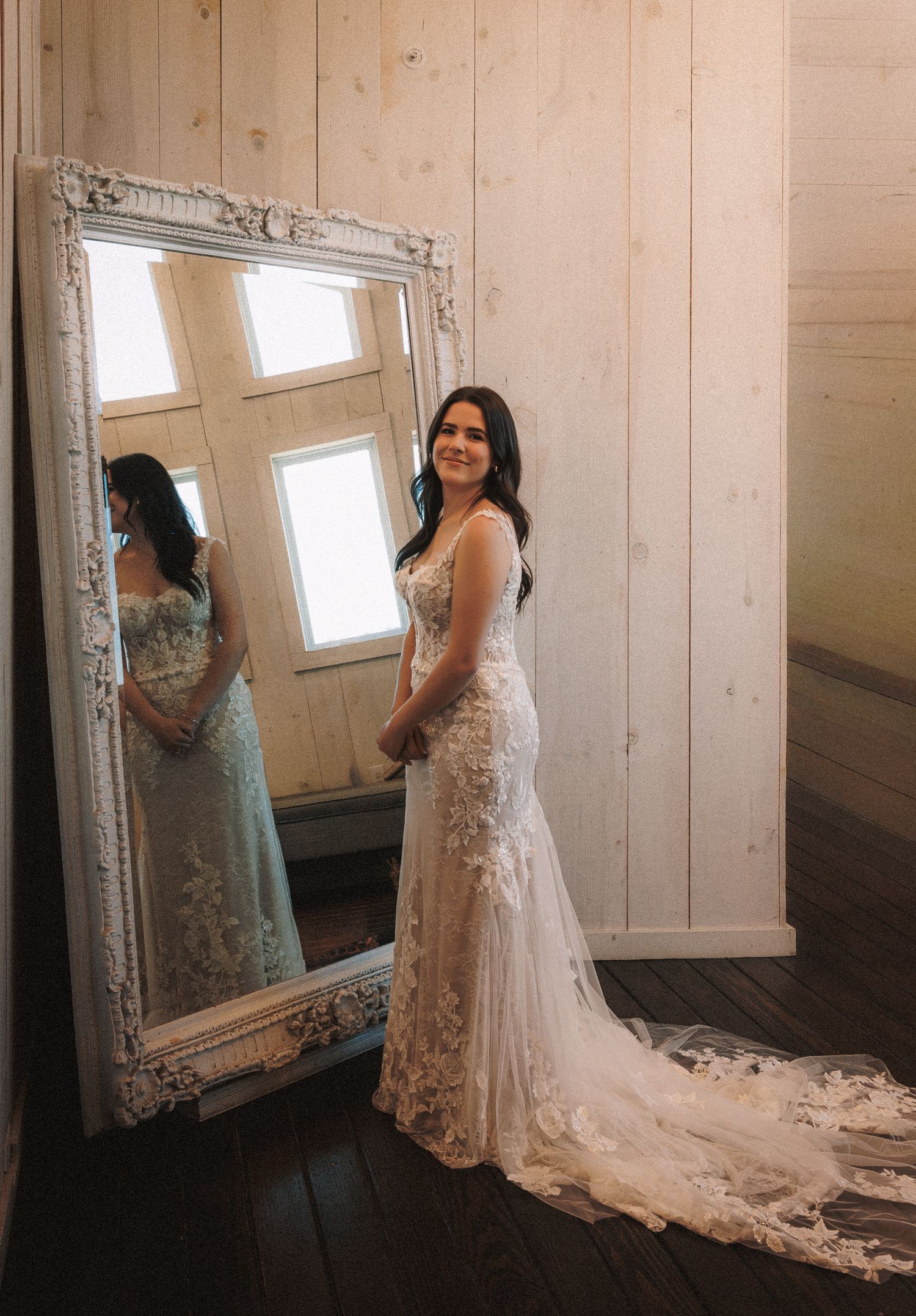 Bride in fitted lace wedding gown with train poses beside ornate white mirror in rustic wood-paneled room.