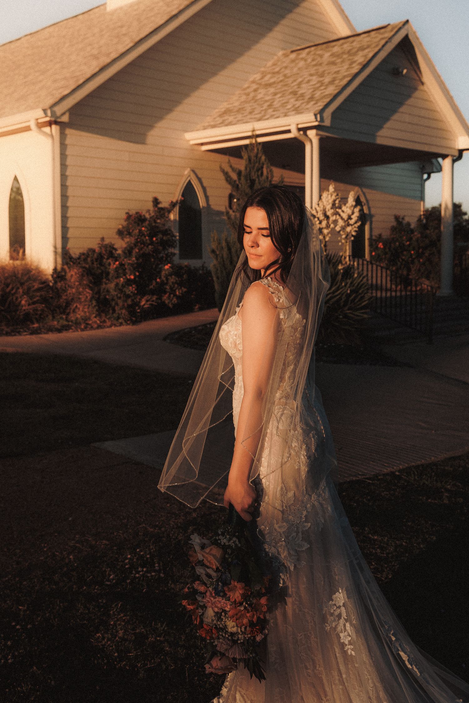 Bride in lace gown and veil holds bouquet while standing before white chapel in golden sunset light.