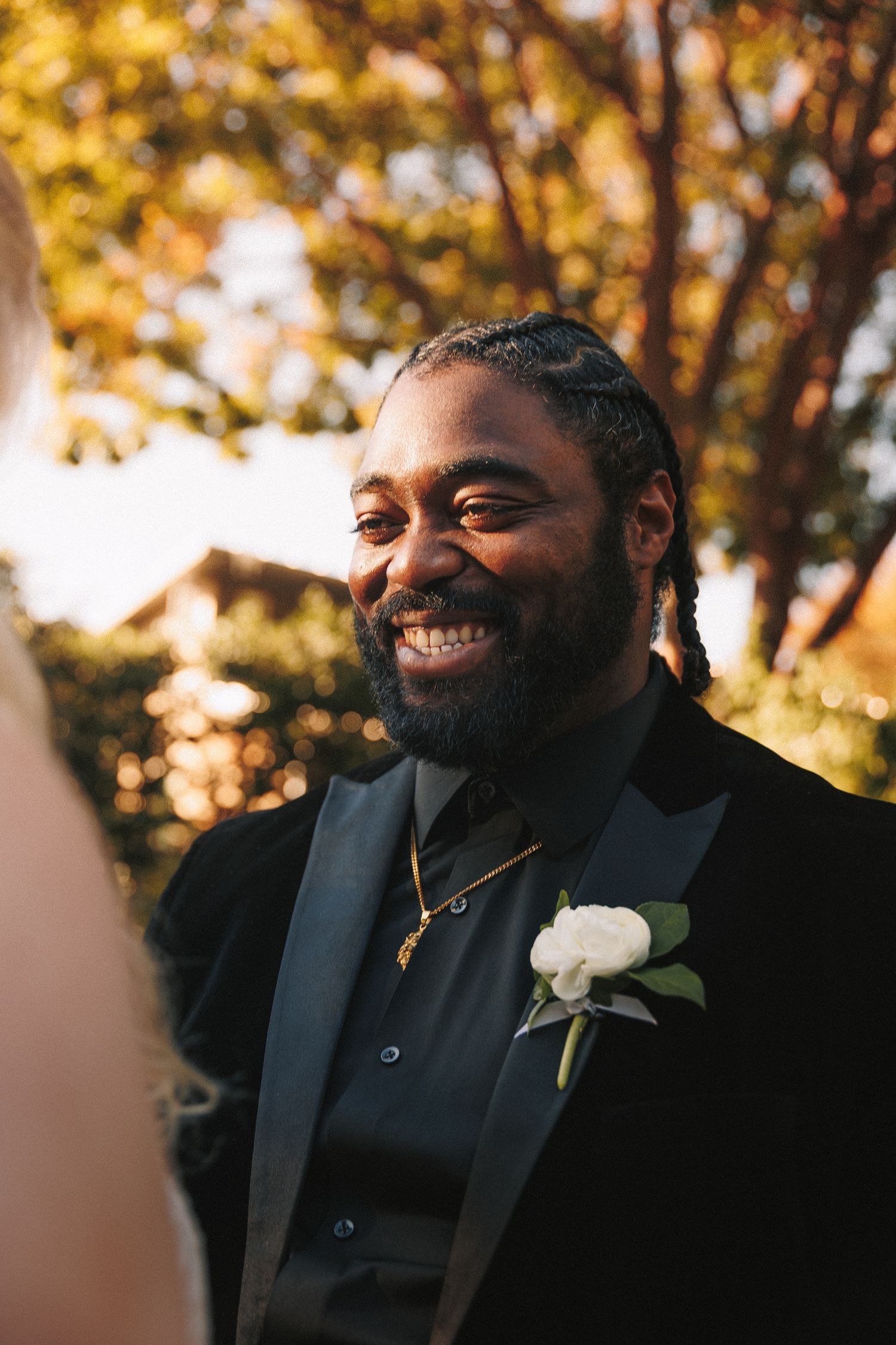Joyful groom in black tuxedo with white boutonniere smiles warmly during outdoor wedding ceremony.