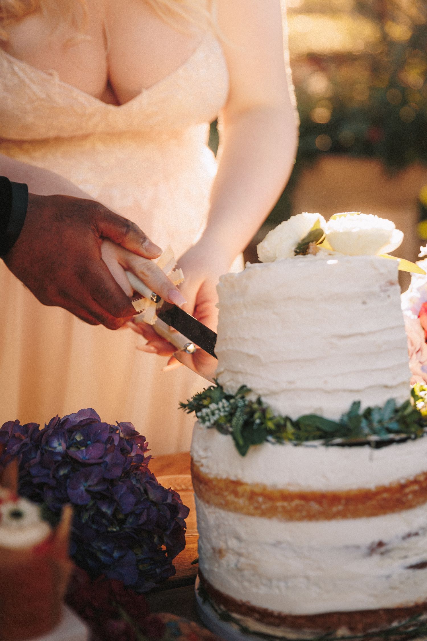 Newlyweds cut a three-tier naked wedding cake adorned with white flowers and greenery at reception.