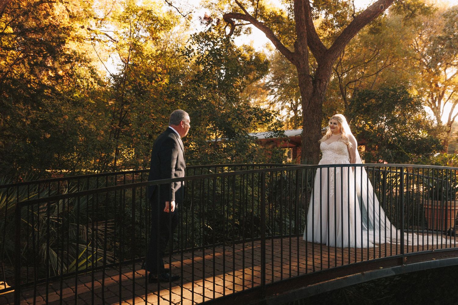 Bride in white gown shares first look moment with father on garden bridge at golden hour.