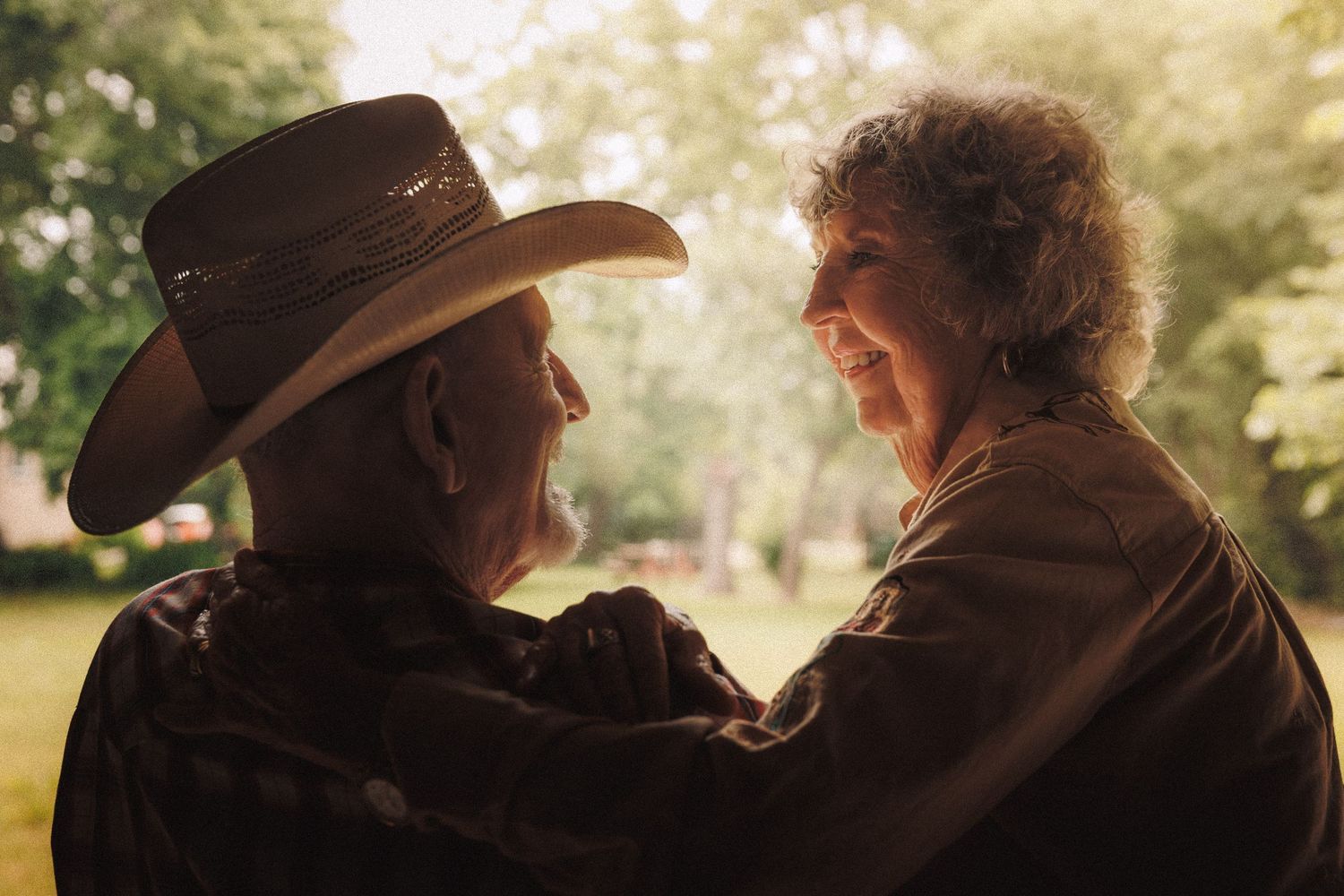 Two people wearing brown cowboy hats sitting closely together outdoors with sunlit trees in the background.