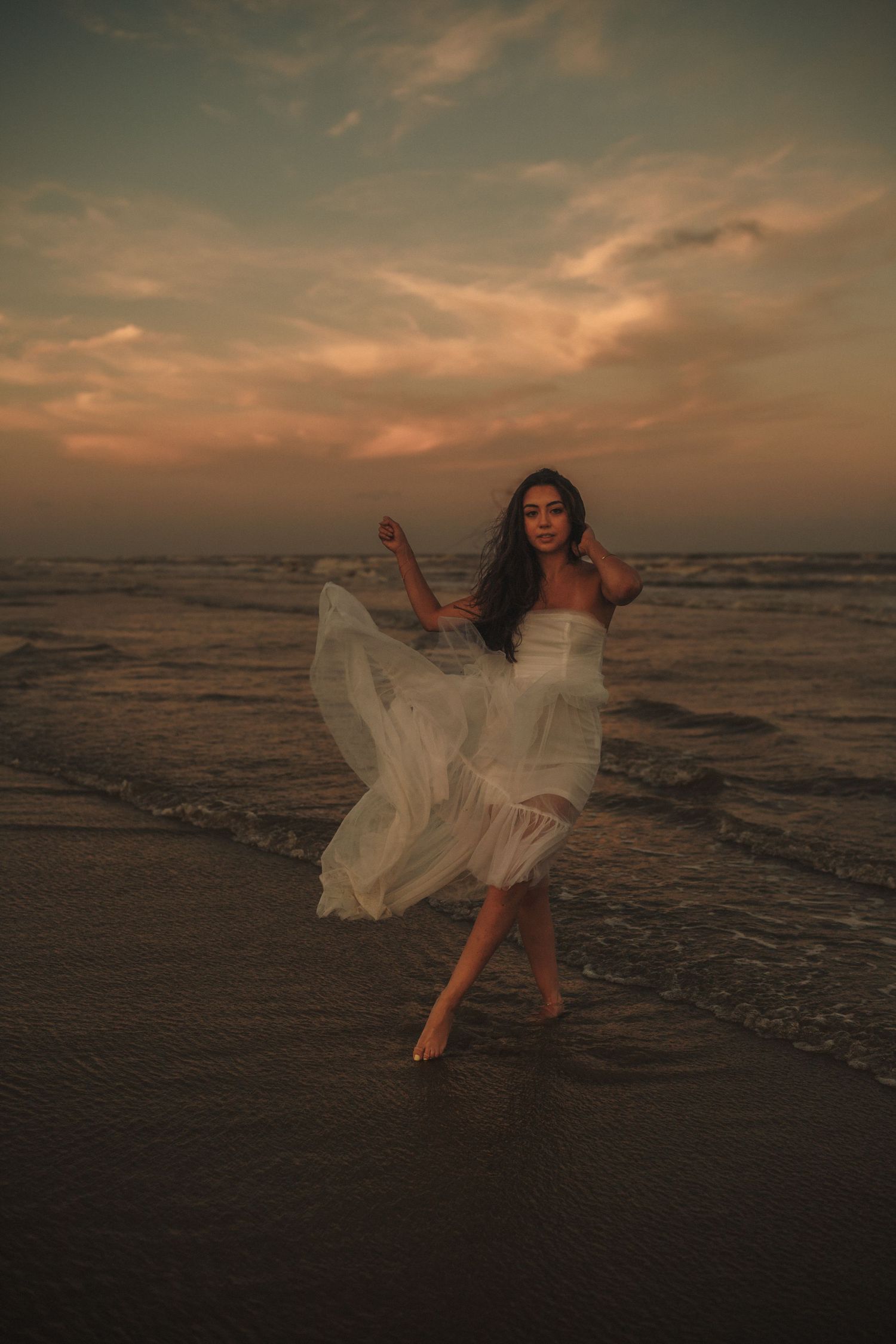 A flowing white dress catches the ocean breeze at sunset as a figure dances gracefully along the shoreline.