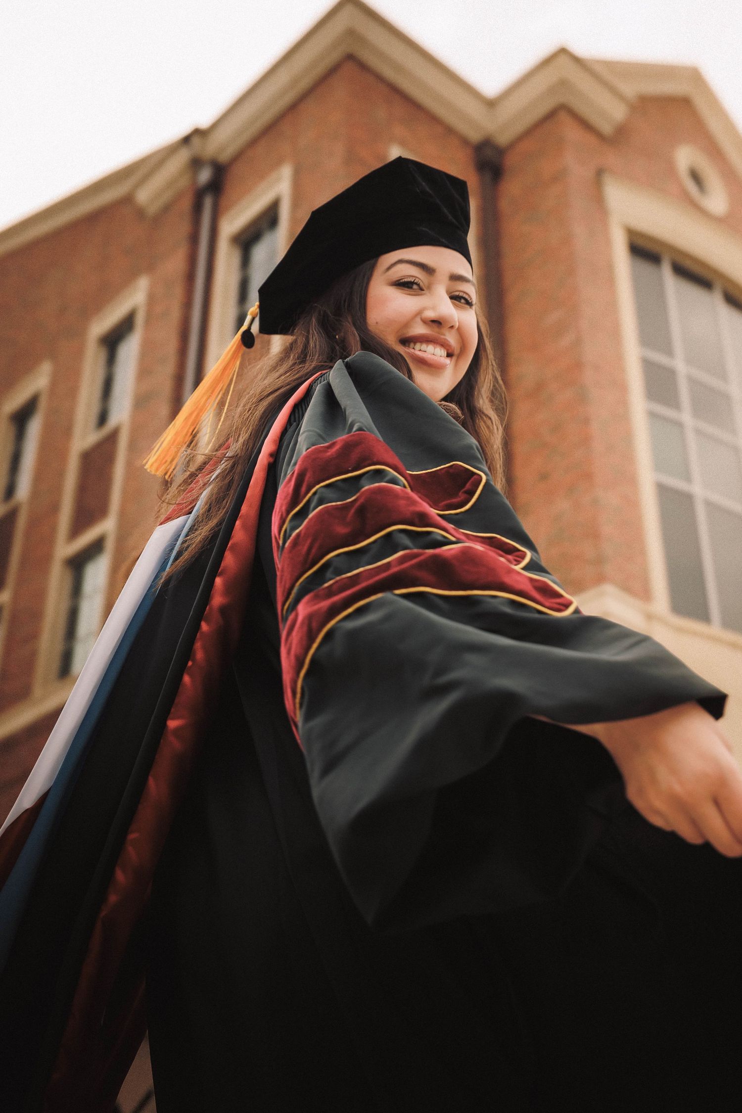 Graduate in black cap and gown smiling during outdoor graduation celebration on campus with brick buildings visible.