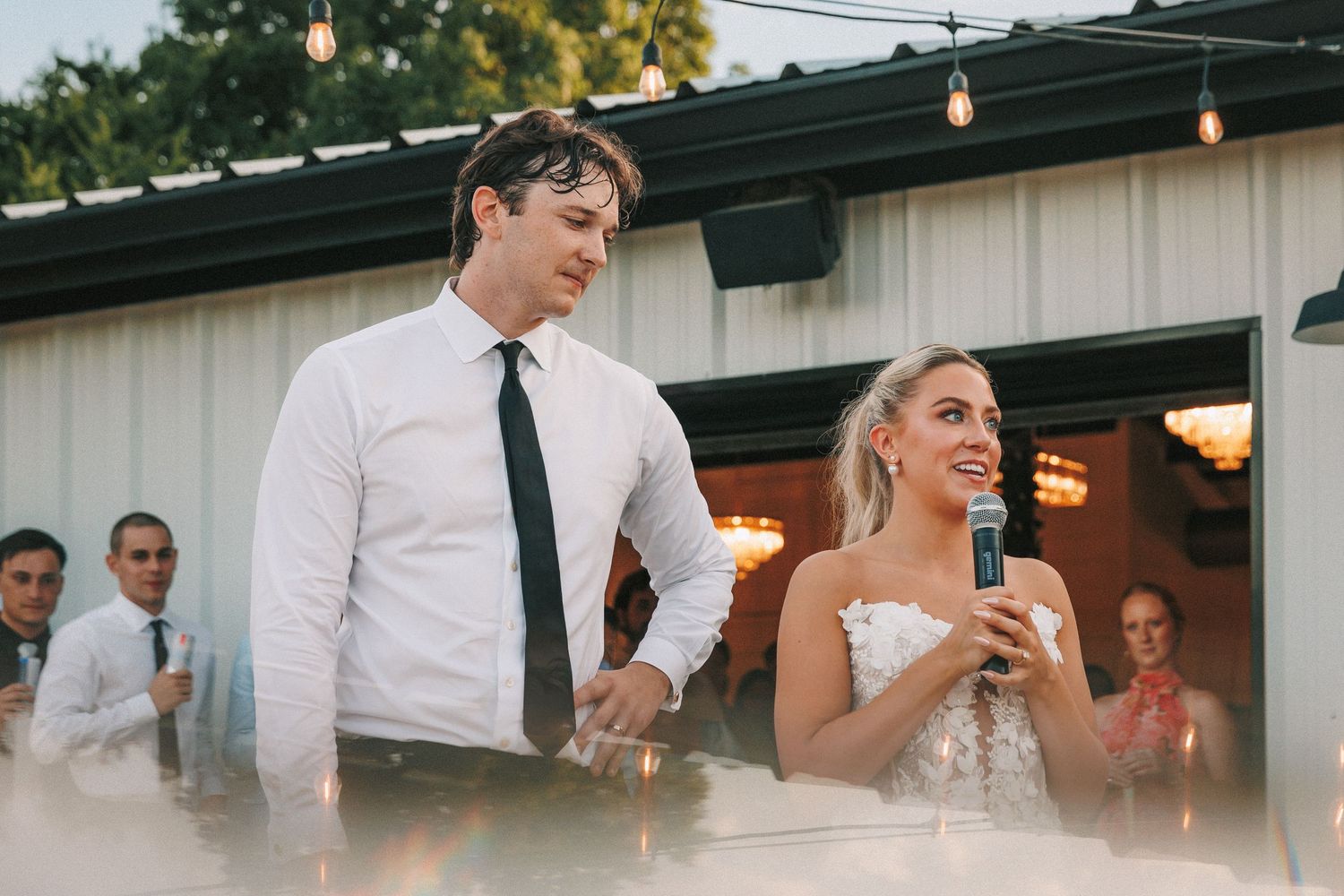 A couple shares a joyful moment during their wedding reception speech under string lights.