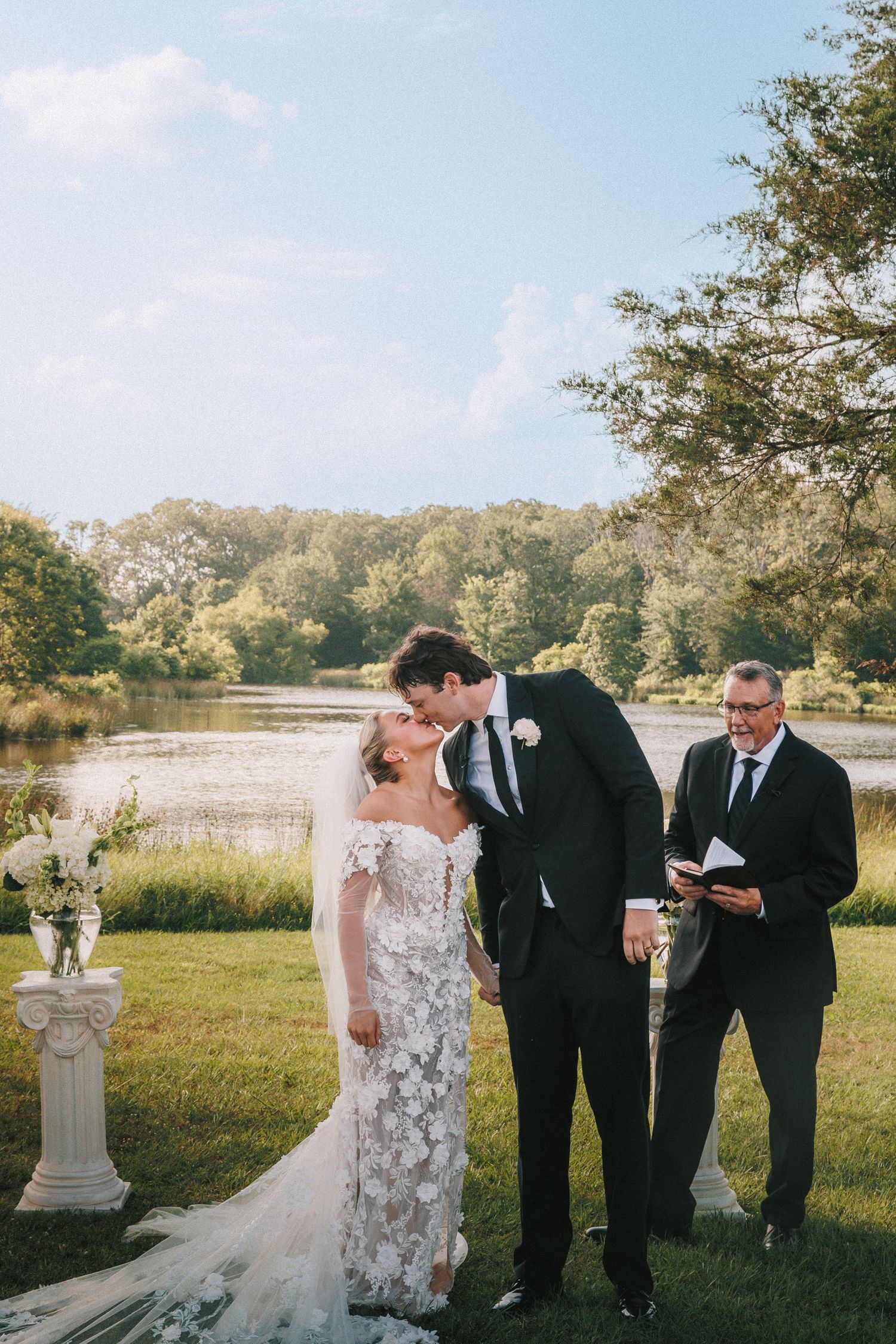 Wedding ceremony being held in a scenic outdoor setting next to a lake with trees in the background.
