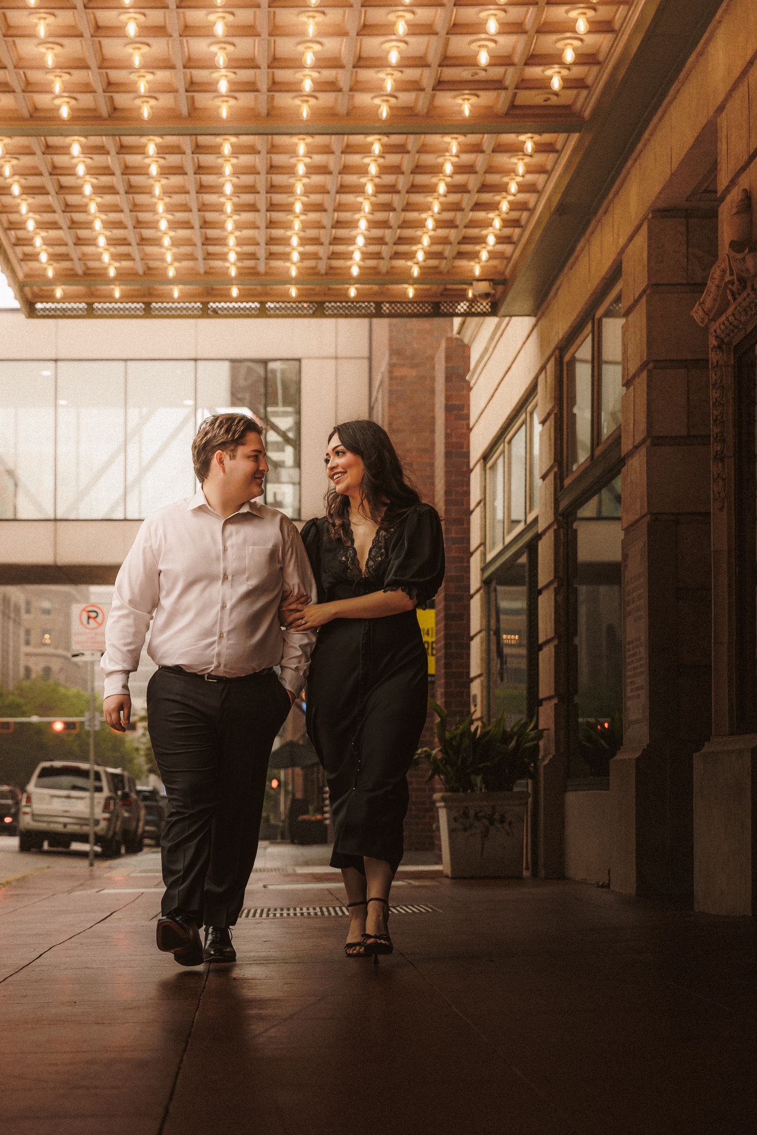 A happy couple walks arm in arm beneath a glowing marquee lights on a city sidewalk.