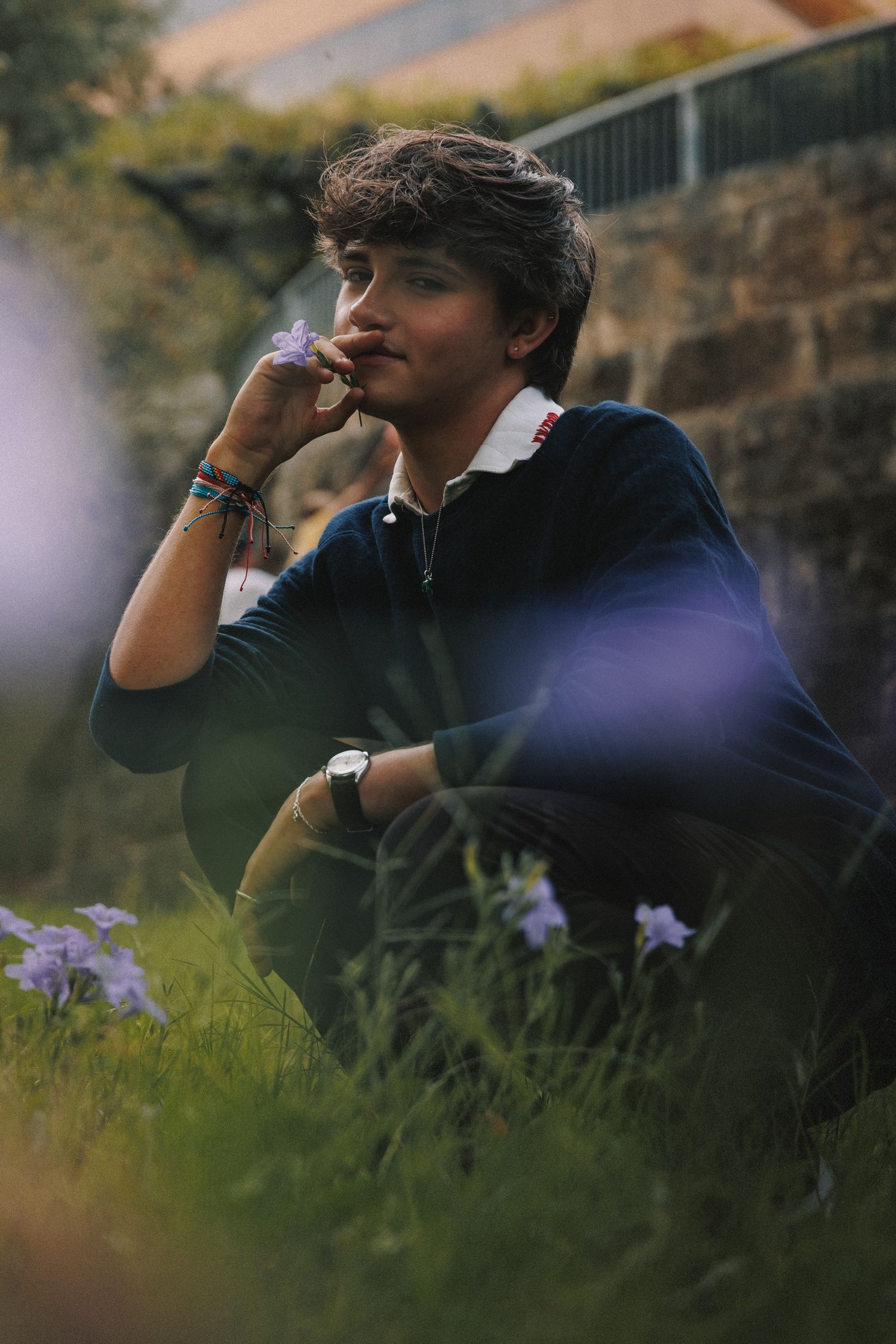 A person sits thoughtfully in a garden with purple flowers, wearing a dark sweater against a stone wall background.