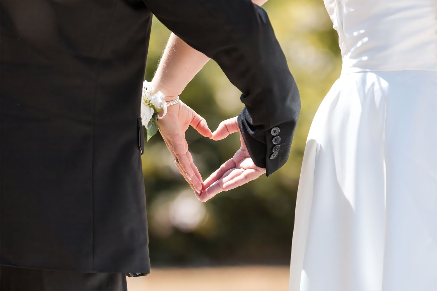 Wedding couple holding hands forming heart shape, bride in white gown and groom in black suit outdoors.