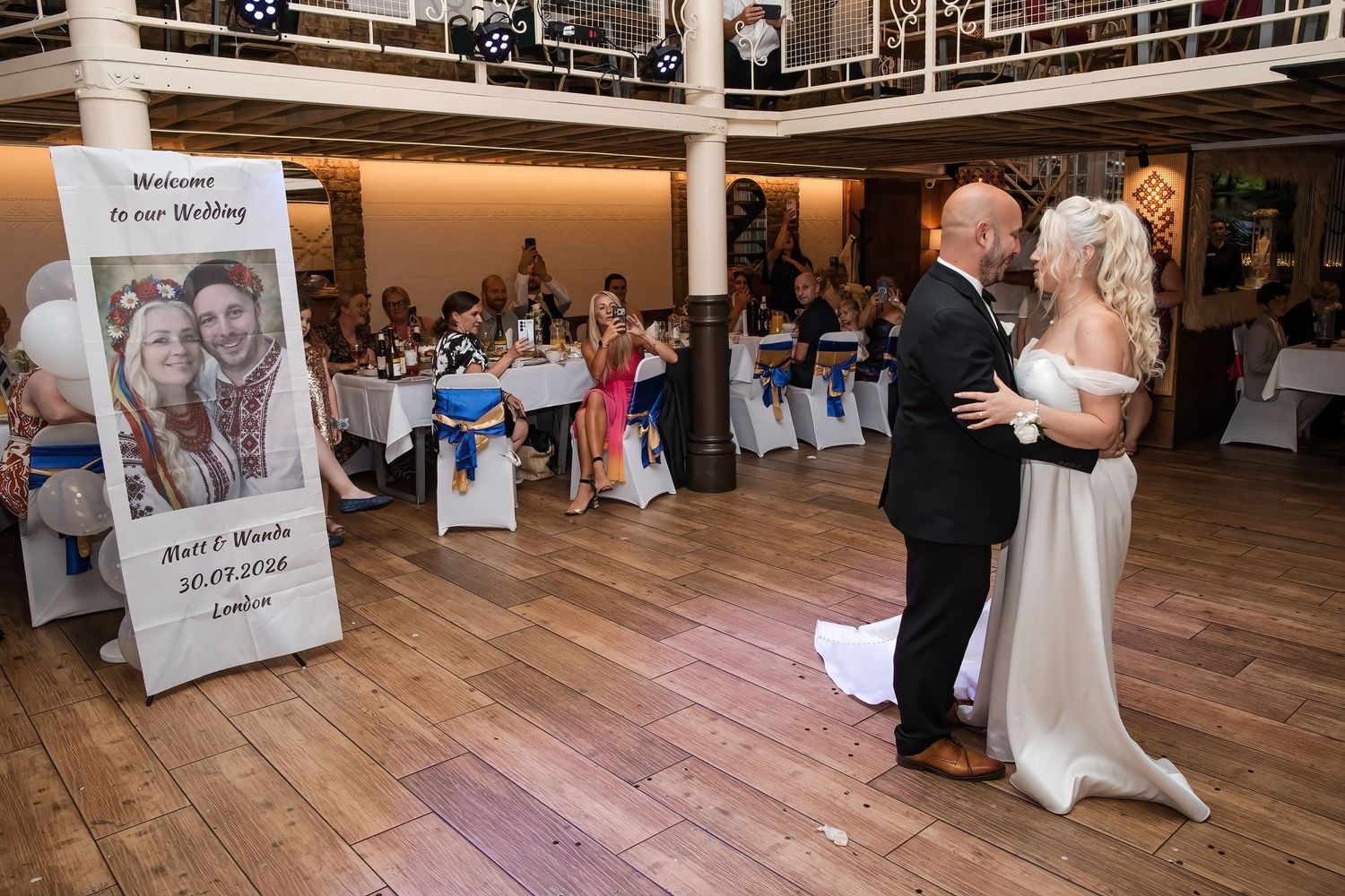 Newlyweds share first dance at wedding reception beside personalized welcome sign on wooden dance floor.