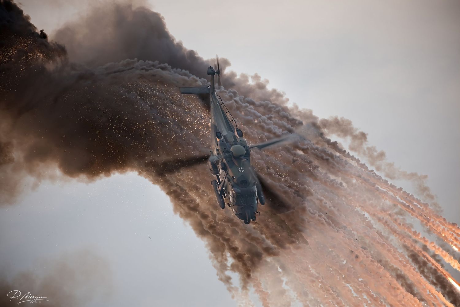 A military helicopter descends through a cloud of golden flare smoke during an airshow.