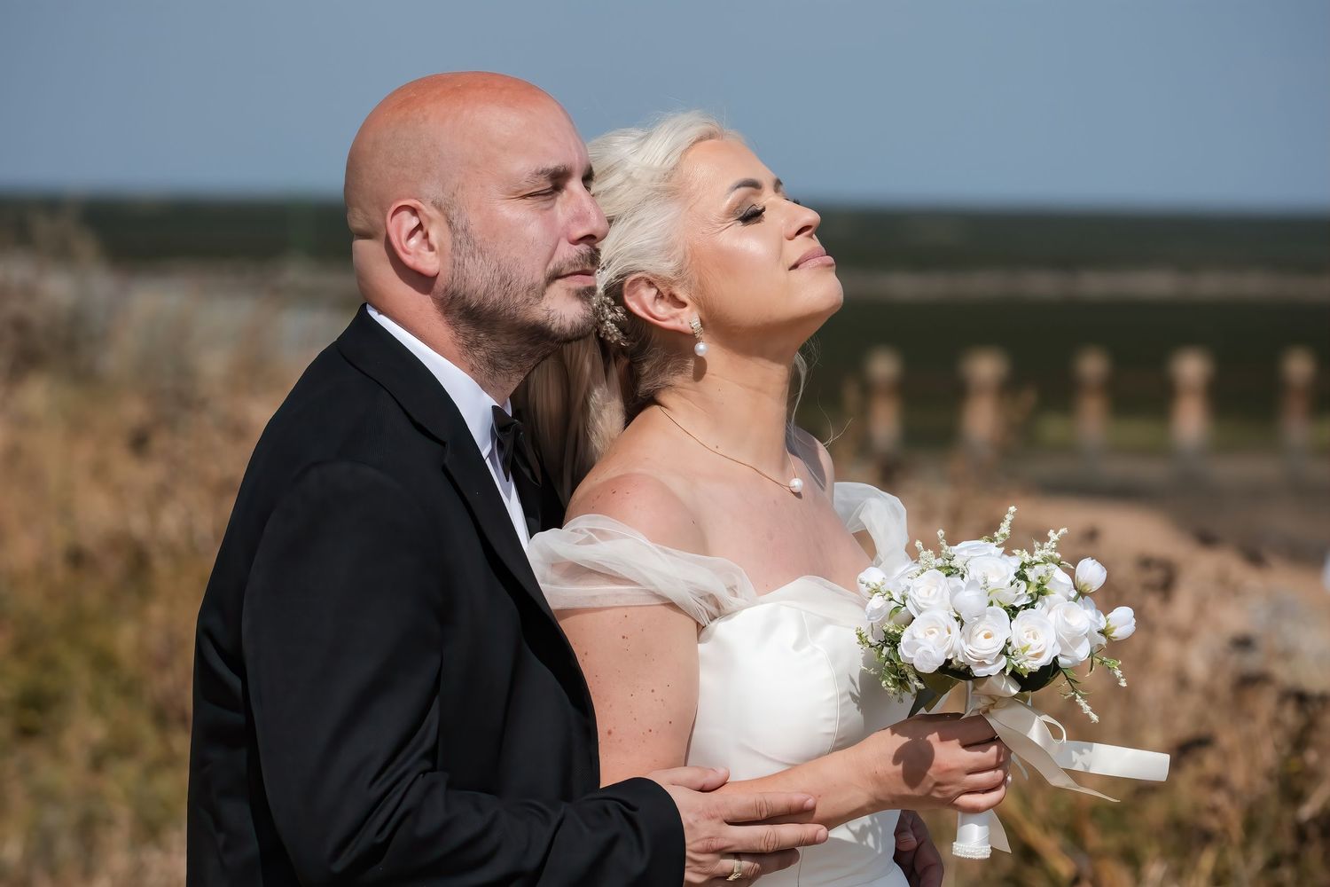 Groom embraces bride from behind as she looks up dreamily, holding white bouquet outdoors.