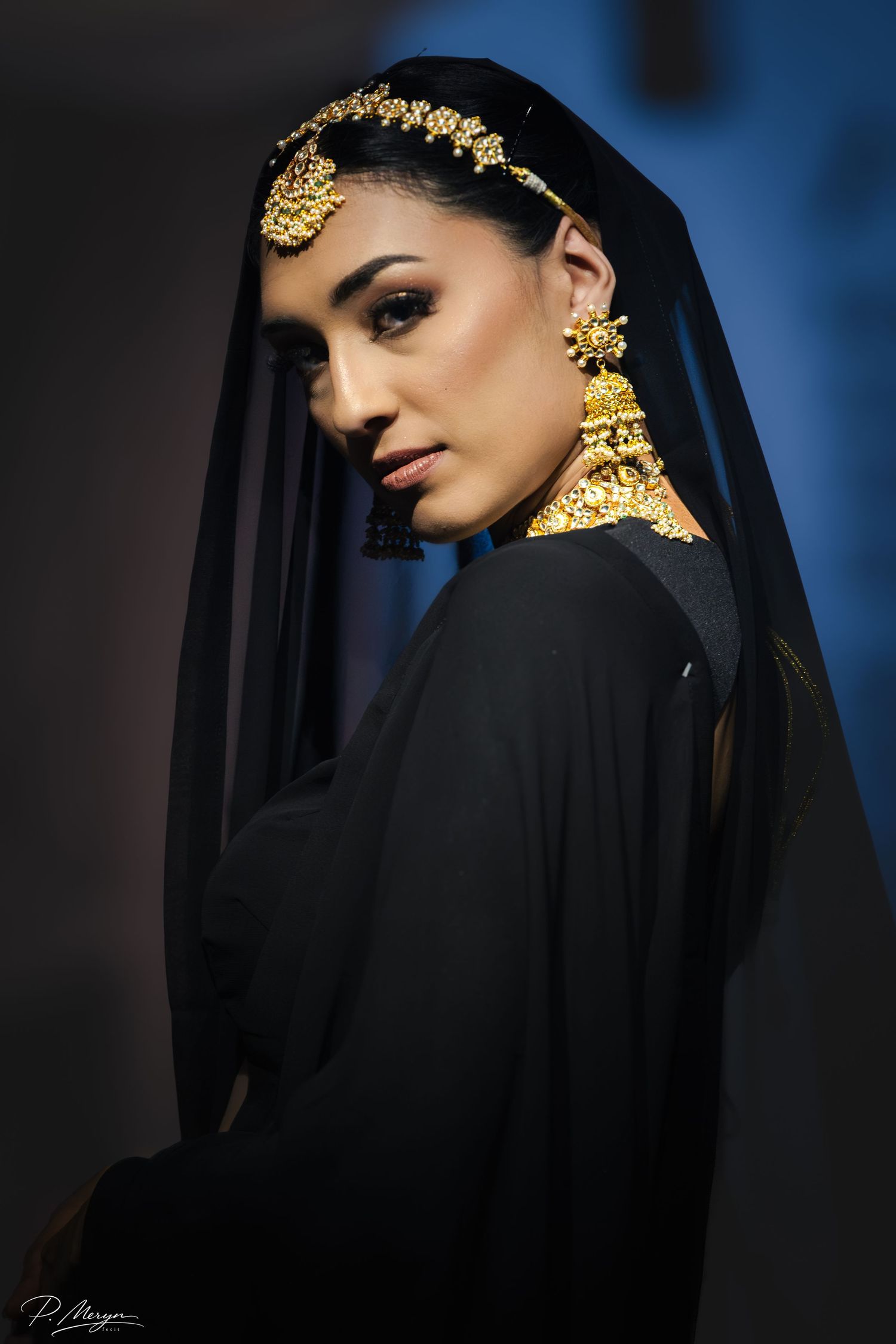 Model wearing black abaya with gold jewelry and headpiece poses elegantly at fashion show.