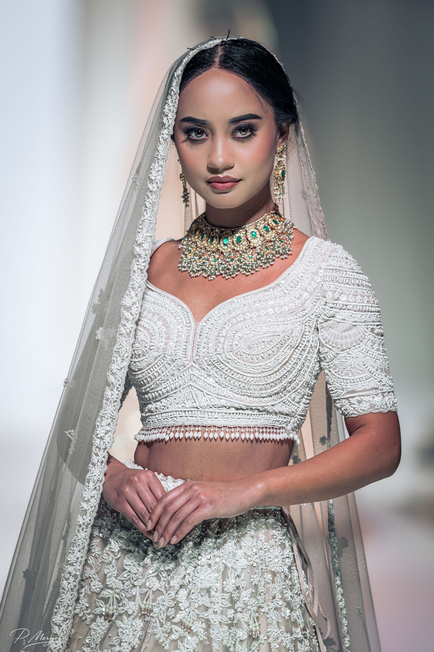 Elegant bride in white embroidered lehenga with emerald choker necklace and embellished veil at Indian wedding.