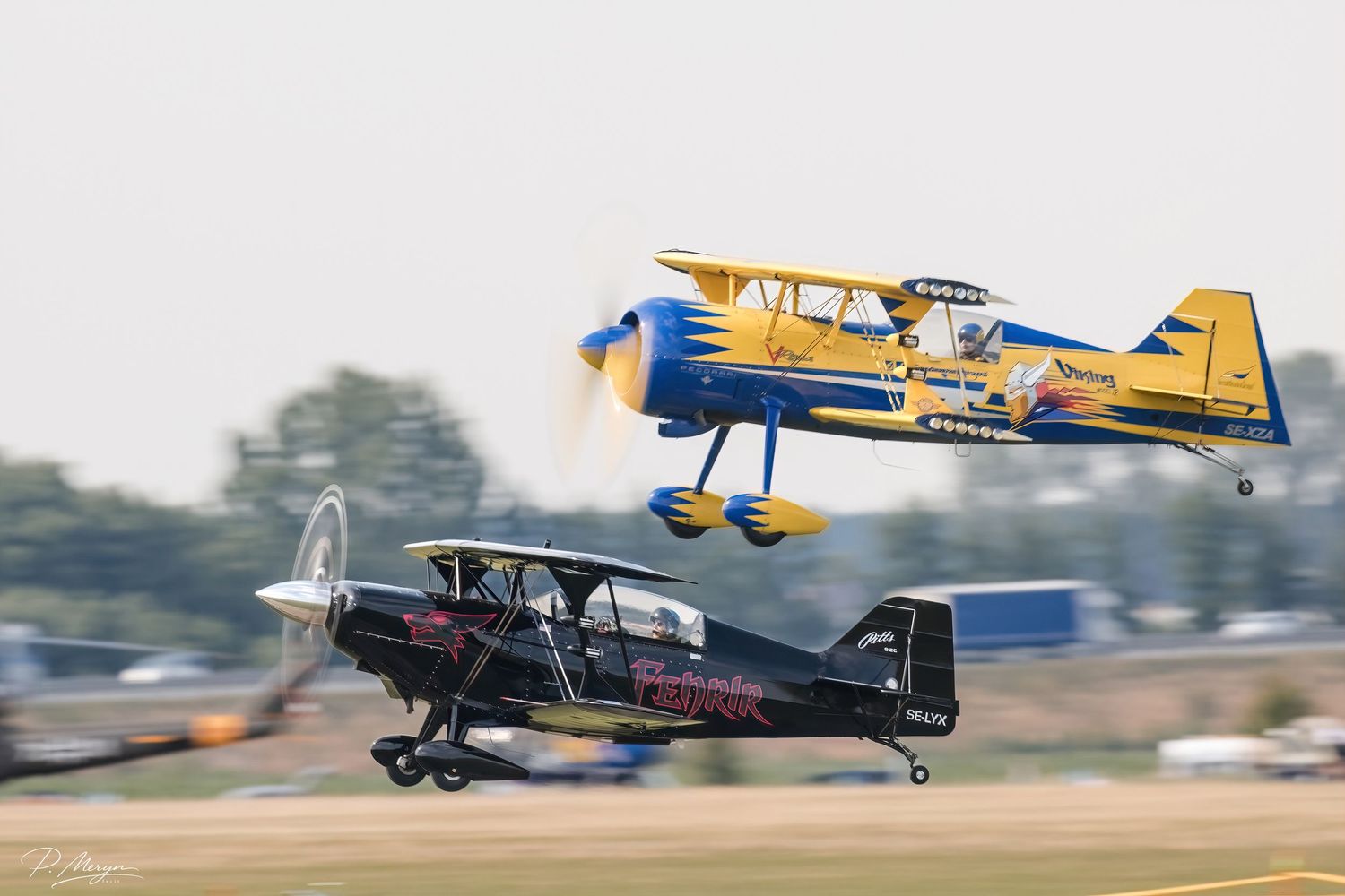 Two colorful Red Bull aerobatic biplanes performing close formation low-level display at summer airshow.