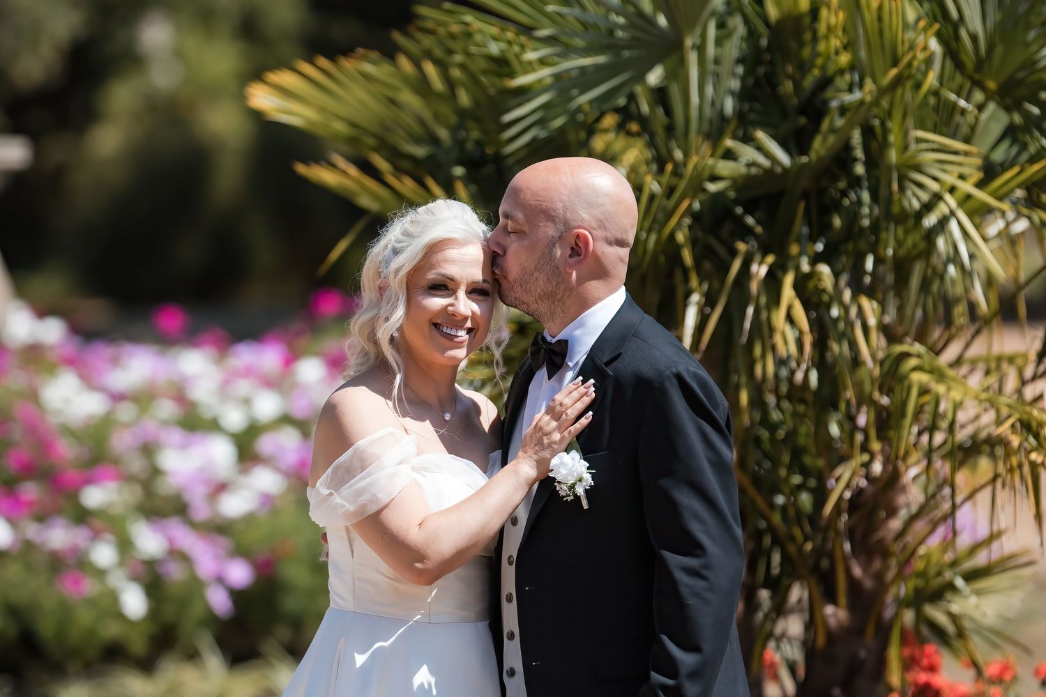 Groom in black tuxedo kissing brides cheek as she smiles joyfully among colorful garden flowers.