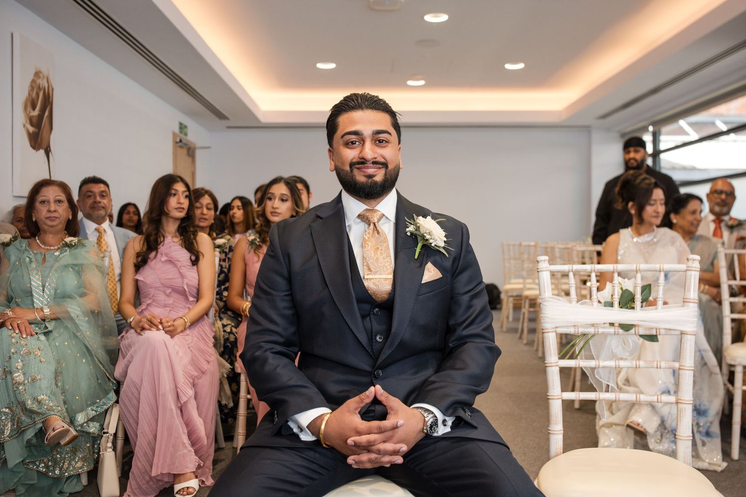 Smiling groom in navy suit with floral boutonniere sits at wedding ceremony awaiting his bride.