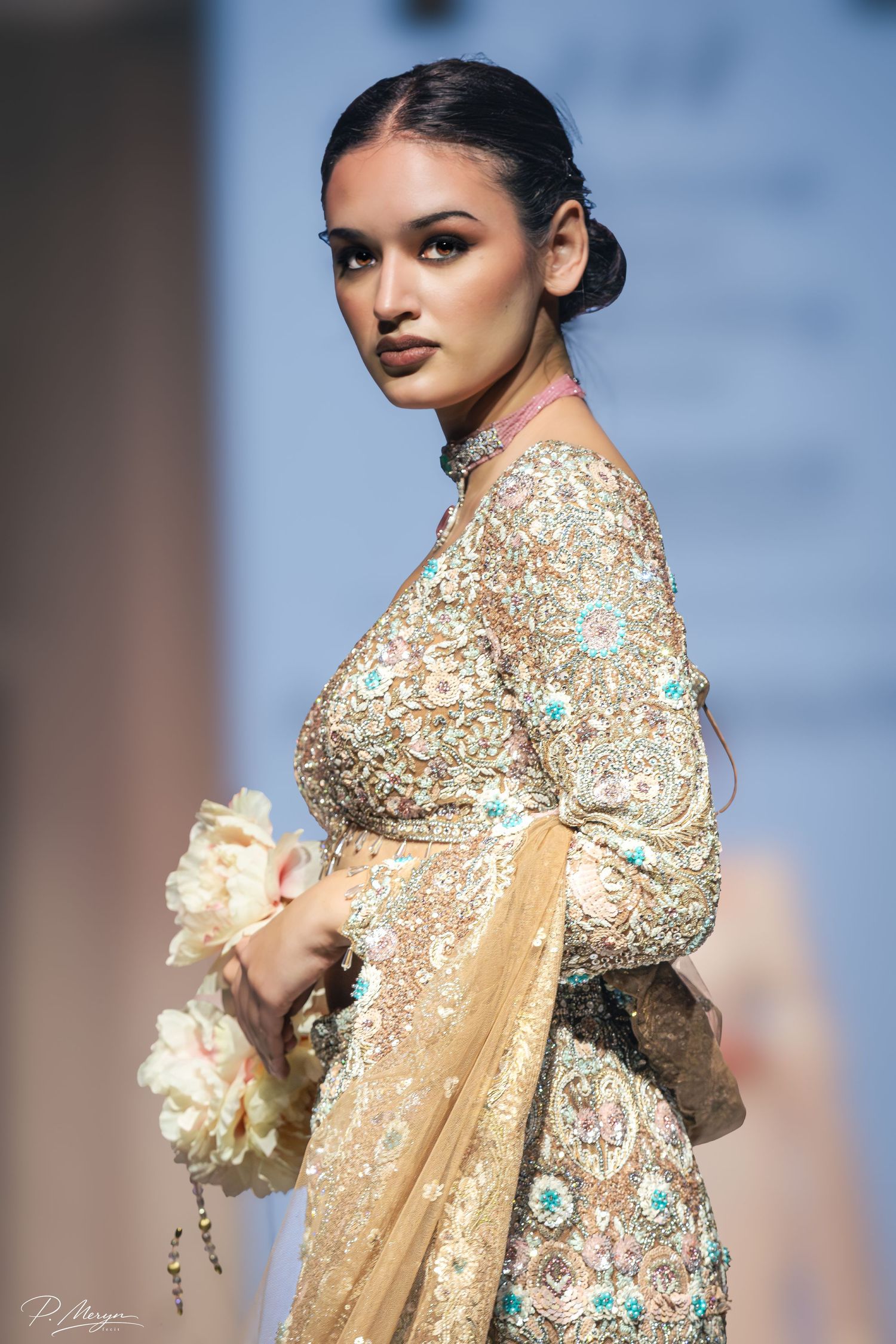 Model in gold embroidered saree with turquoise floral details holds white flowers on fashion runway.