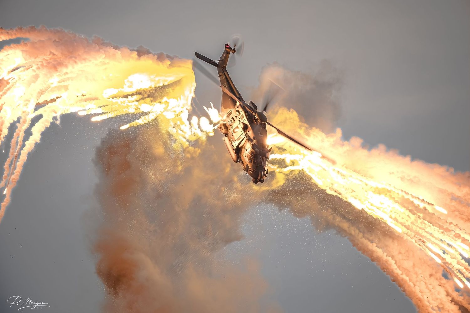 A military helicopter releases dramatic flares creating fiery trails against a grey sky.