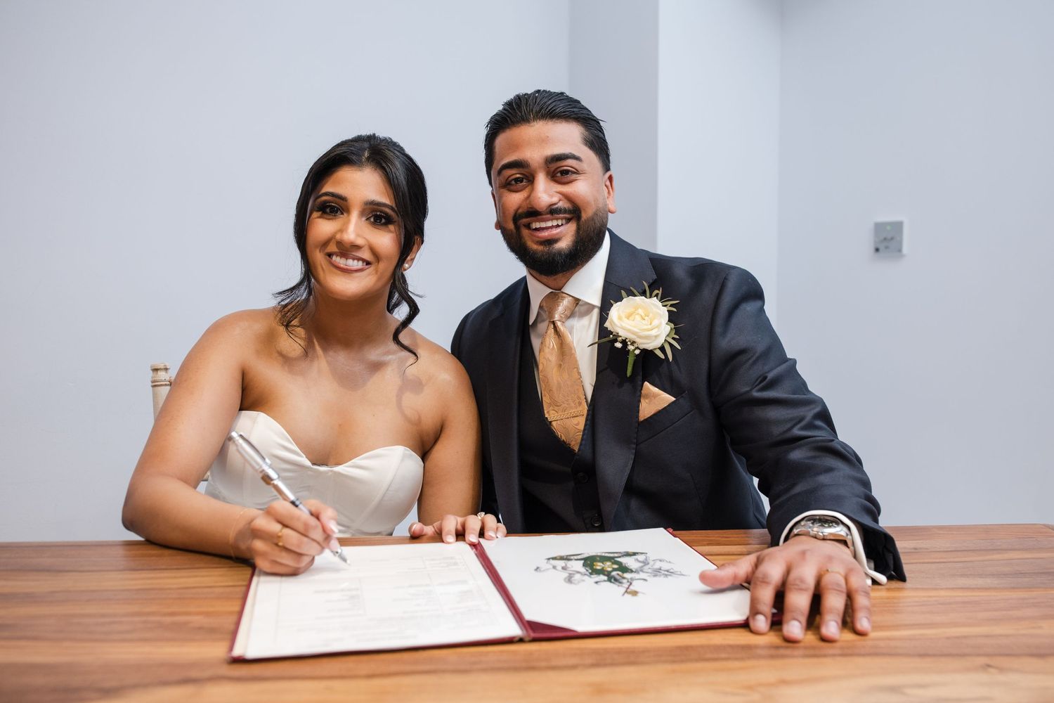 Smiling newlyweds signing wedding register at table with groom in suit and boutonniere.
