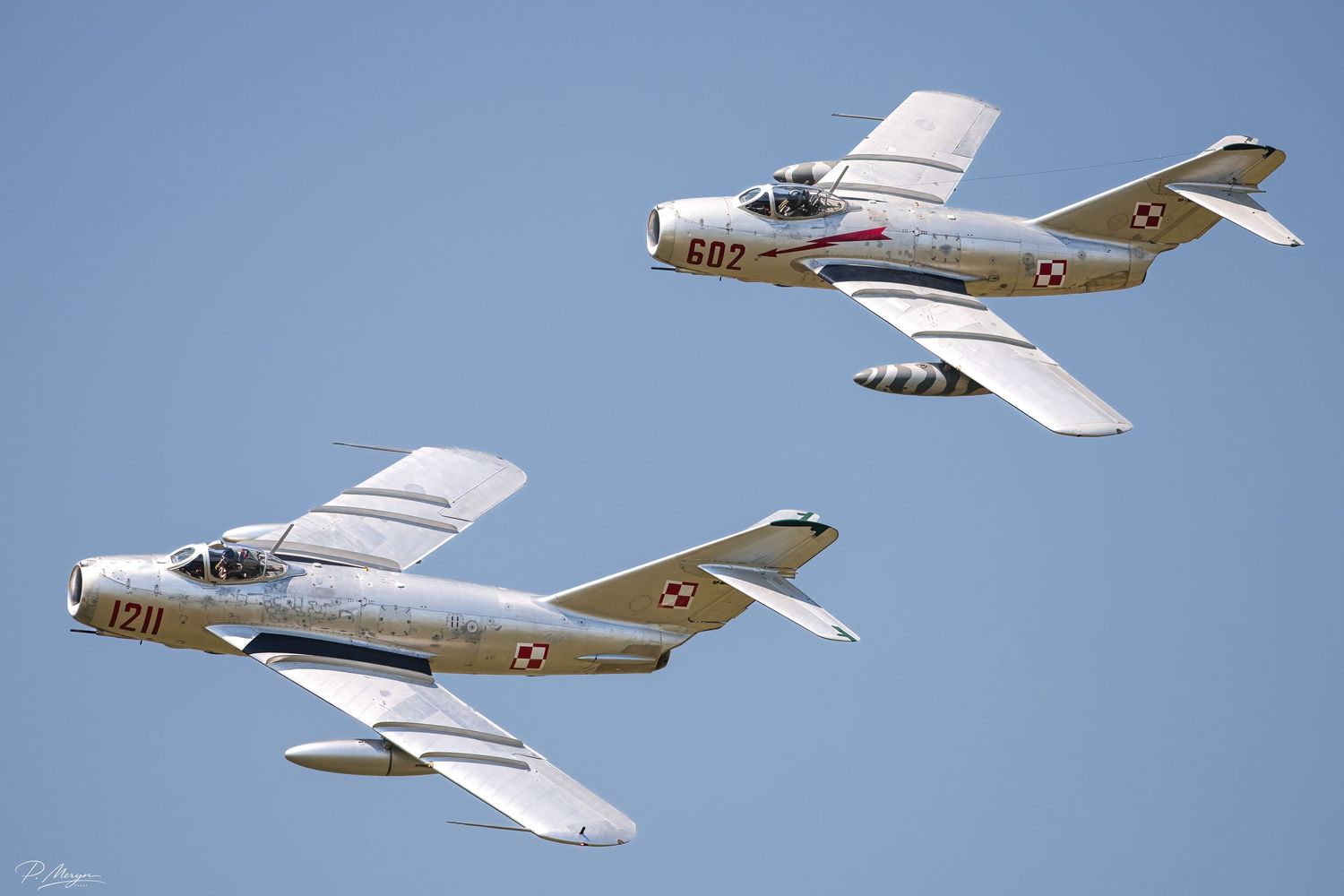 Two Polish MiG-15 vintage jet fighters in silver and white livery flying in formation.
