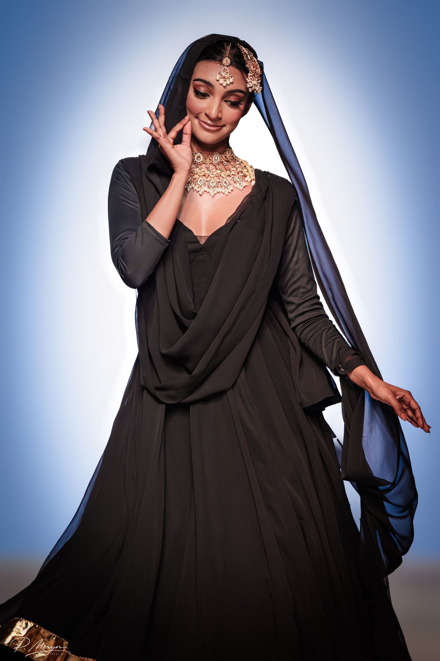Model in black lehenga with gold jewelry and blue dupatta poses gracefully on fashion runway.