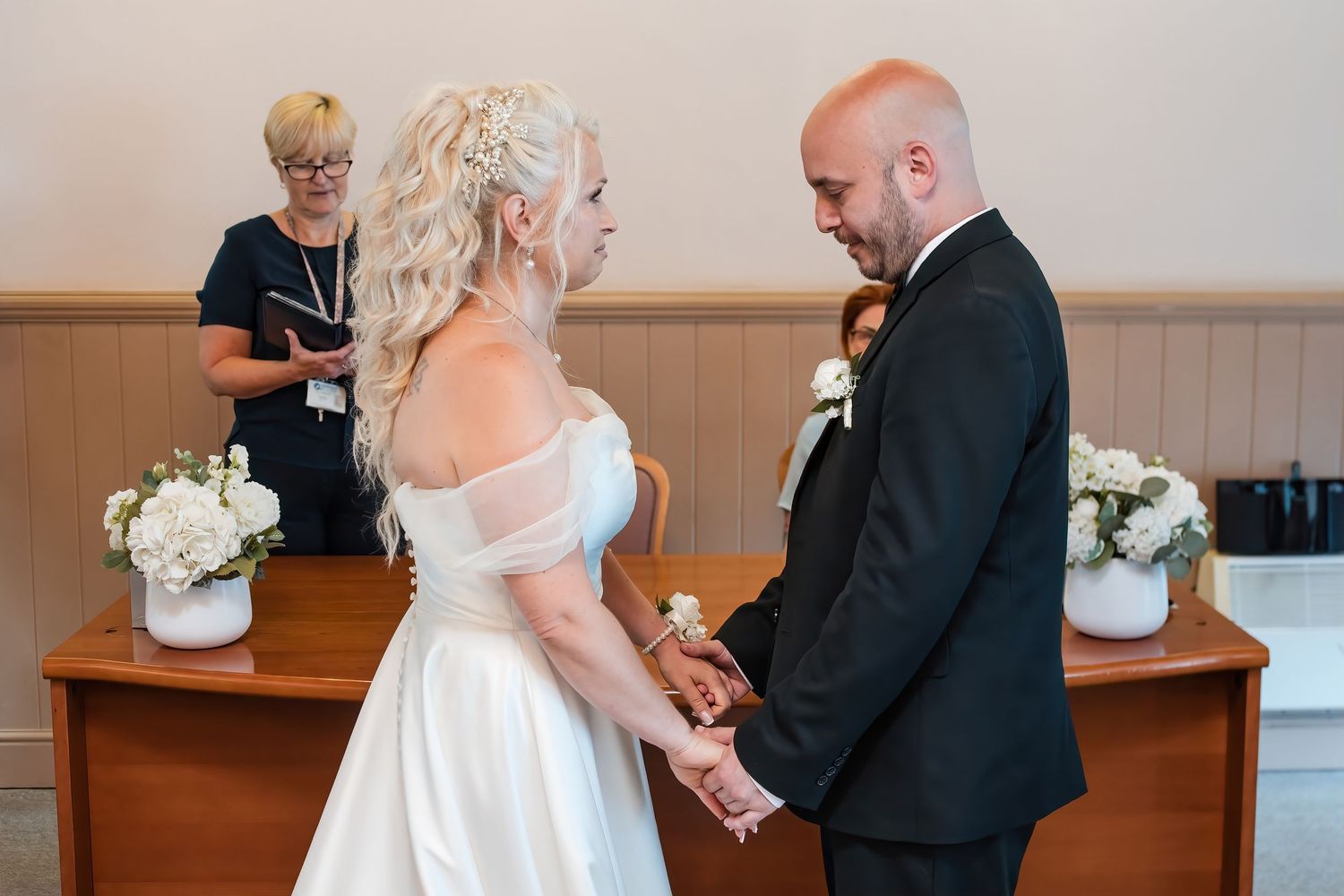 Bride and groom hold hands facing each other during wedding ceremony with officiant behind.