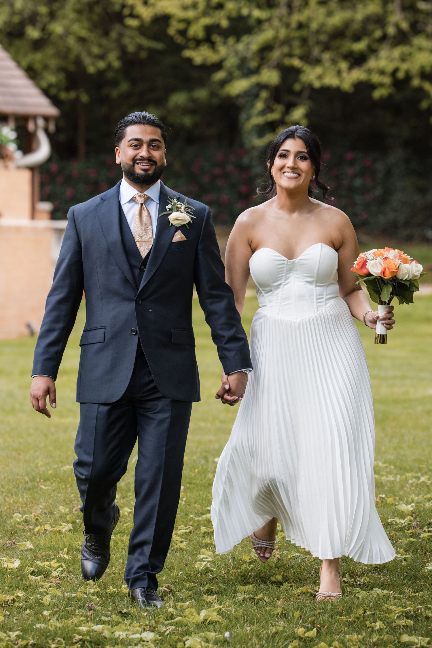 Smiling bride in white pleated gown holds bouquet while walking hand-in-hand with groom in navy suit.
