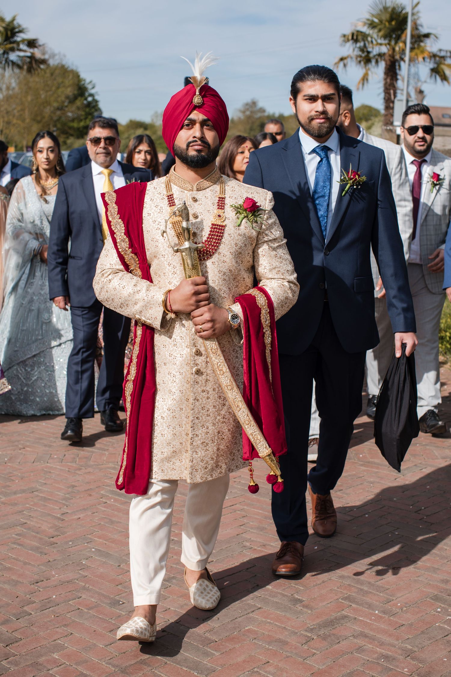 Sikh groom in traditional cream sherwani and red turban leads baraat procession with guests outdoors.