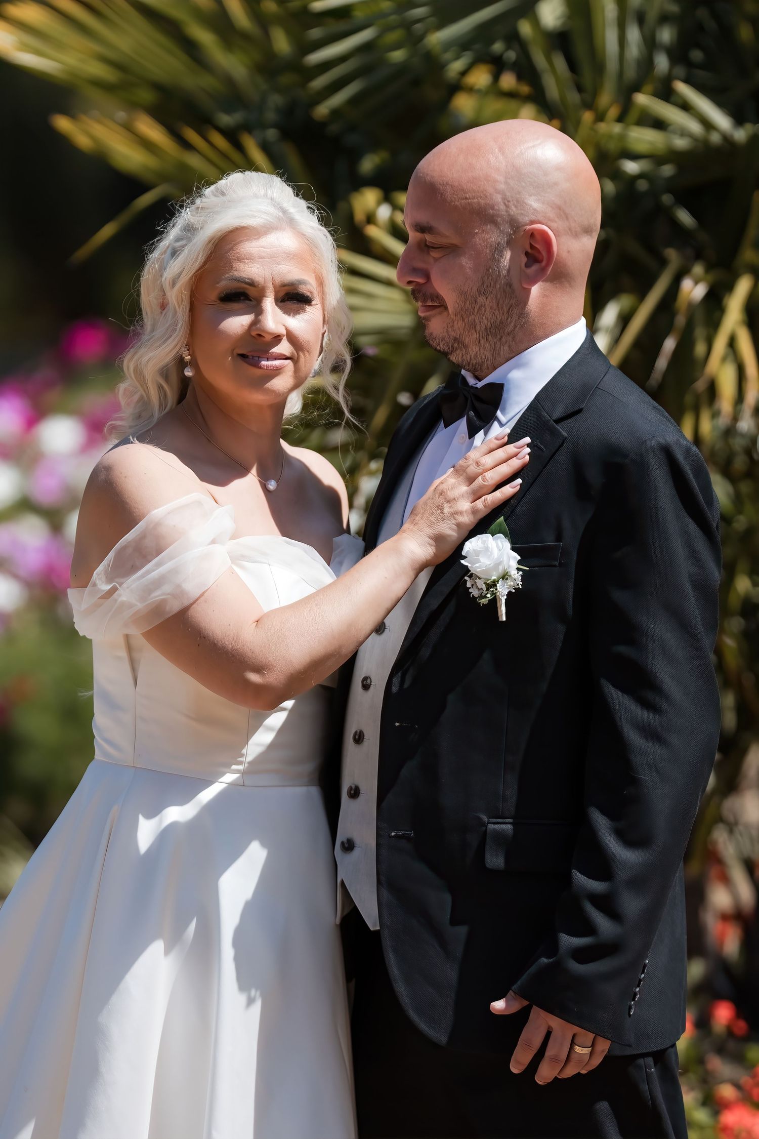 Elegant bride in off-shoulder white gown places hand on grooms chest during sunny outdoor wedding ceremony.