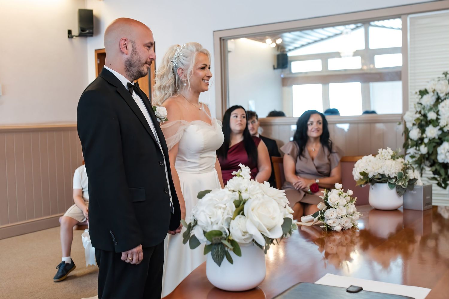 Bride and groom stand at altar during wedding ceremony with white floral arrangements nearby.