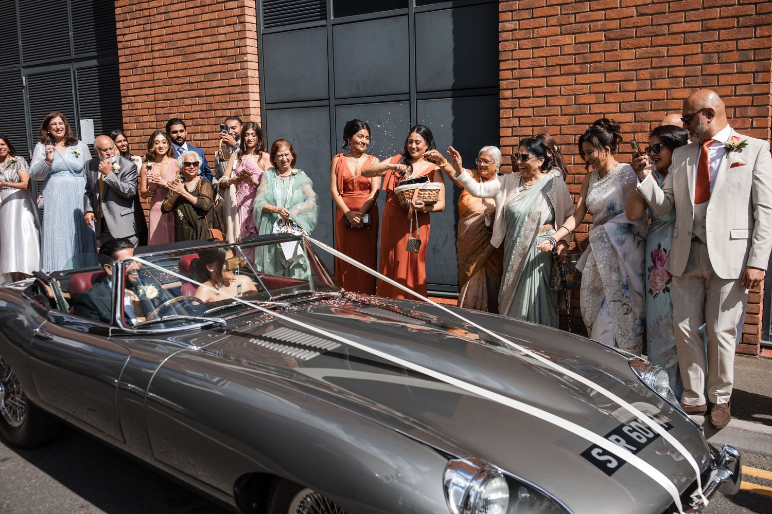 Wedding guests cheering as bride and groom depart in a classic silver Jaguar convertible outside brick building.